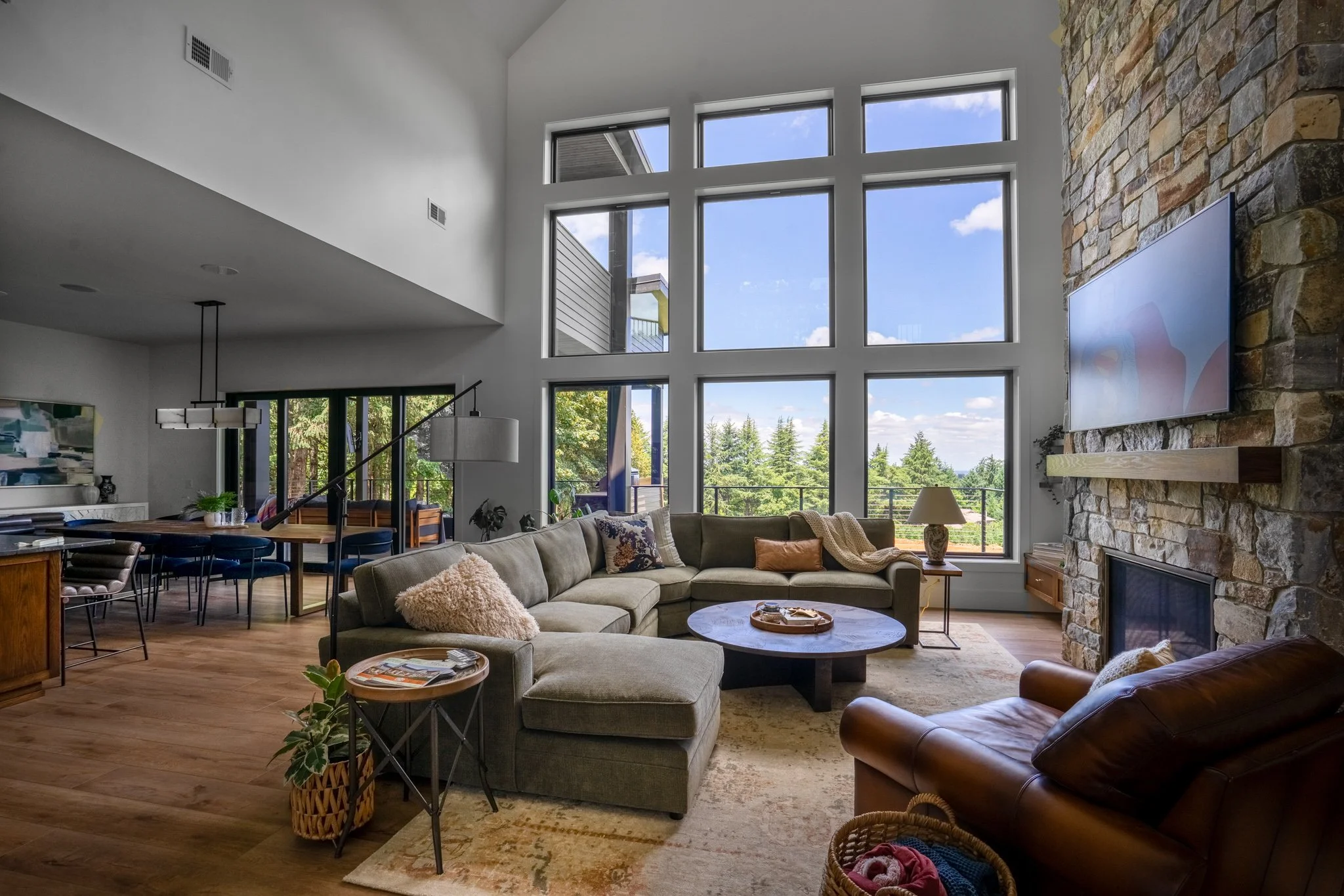 A spacious living room with large grid windows showing trees and a blue sky, a stone fireplace with a mounted TV, beige sofas with pillows and blankets, a round coffee table, wooden flooring, and a dining area in the background.