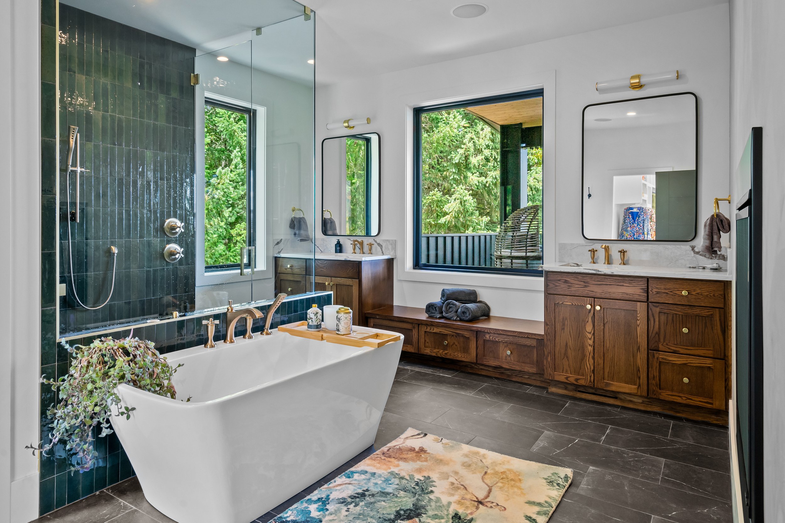 Modern bathroom with a free-standing bathtub, a shower area with dark green tiles, two large windows showing outdoor greenery, wooden vanity with two mirrors, and a colorful rug on the dark tiled floor.