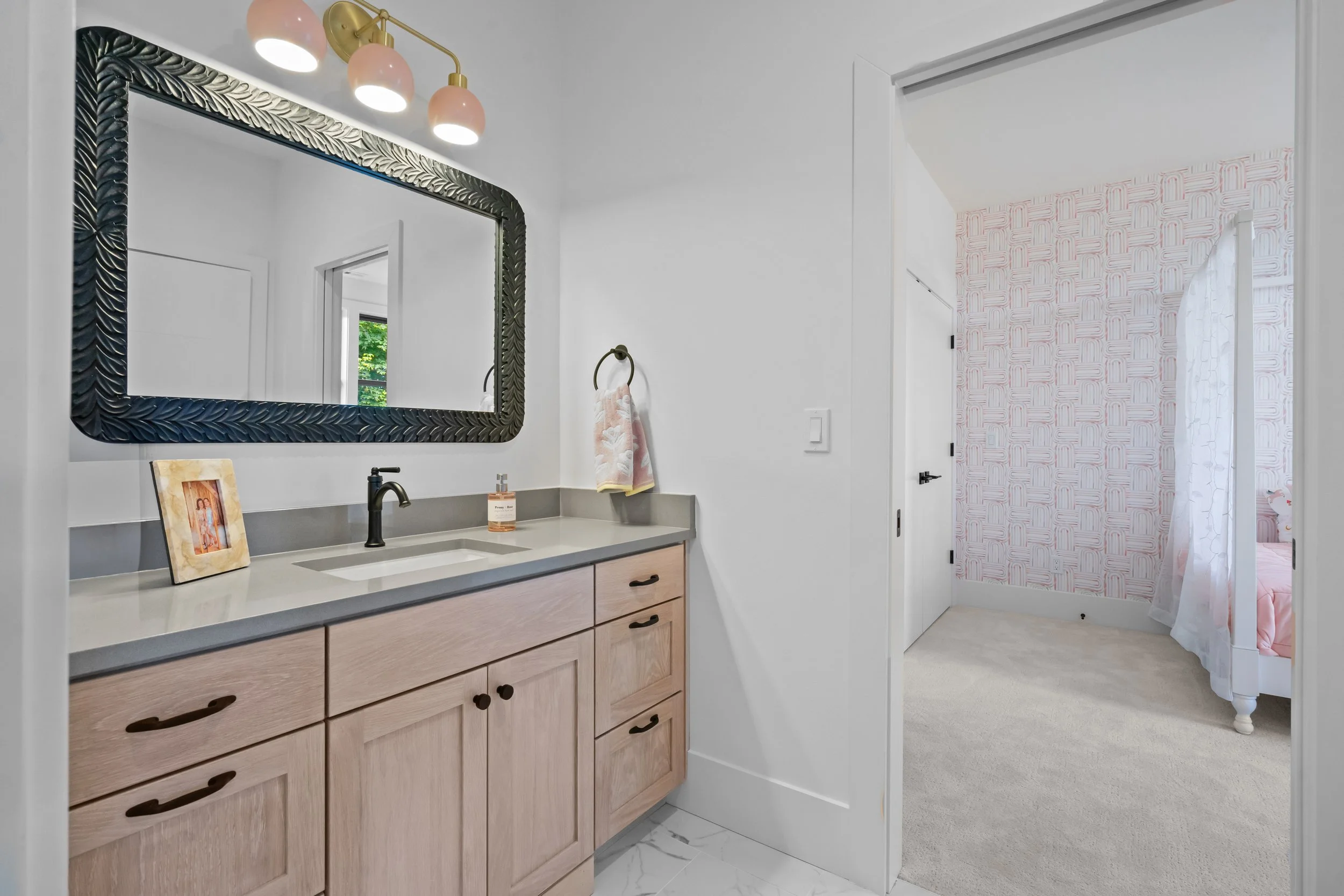 Bathroom vanity with a large framed mirror, three pink pendant lights, a towel ring with a pink and white towel, and part of a doorway leading to a room with pink and white patterned wallpaper and a bed.