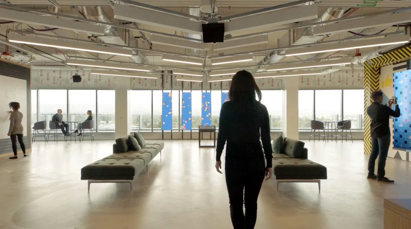 Interior of a modern office or co-working space with large windows, contemporary seating, and people engaging with digital displays.