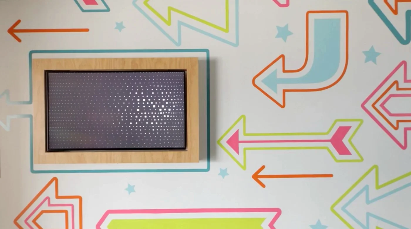 A wall decorated with colorful arrows and small stars, with a framed screen or picture on the left side surrounded by a light wood border.