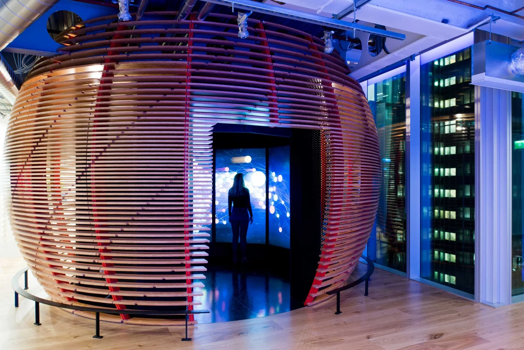 A person standing inside a circular, art installation made of horizontal, wooden slats with red accents, illuminated with purple and blue lighting, located in a modern building with glass walls and wooden flooring.