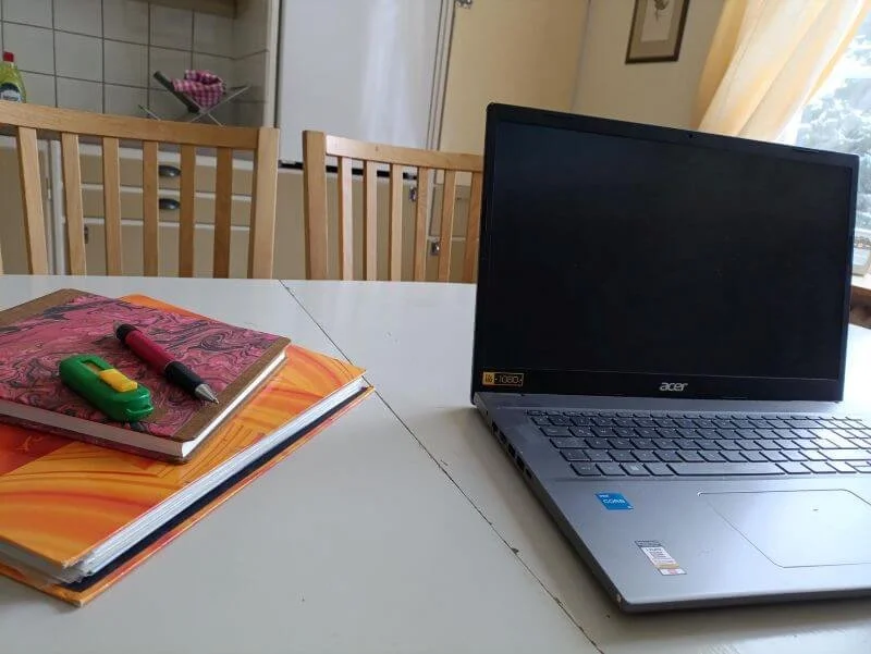 Ein Esstisch mit mehreren Stühlen, auf dem ein Laptop, einige Notizbücher, ein Textmarker und ein Kugelschreiber liegen. Im Hintergrund eine Küche und ein Fenster mit Vorhängen.