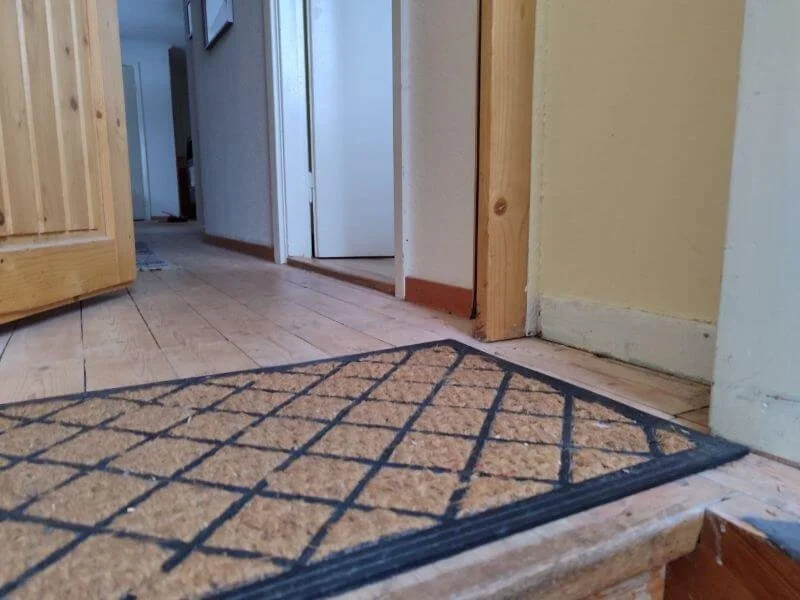 Innenaufnahme eines Hausflurs mit Holzboden, einem Eingangsbereich mit Fußmatte und mehreren Türen.