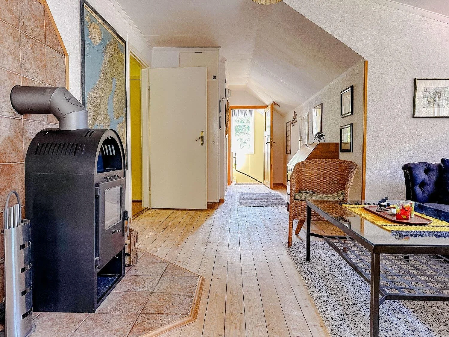 Gemütliches Wohnzimmer mit Holzfußboden, Rattan-Sessel, geschnitztem Holz-Schreibtisch, Fernsehtisch, kunstvollen Bildern an der Wand, Kaminofen, Tür zum Garten, und natürlichem Licht von Fenstern im Hintergrund.