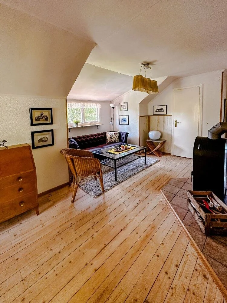 Gemütliches Wohnzimmer mit Holzfußboden, braunem Sofa, Rattanstuhl, Couchtisch, Wandbilder, Stehlampe, große Fenster mit Weißvorhängen, Holzregal, White Vase auf einem kleinen Tisch, Kamin auf der rechten Seite.