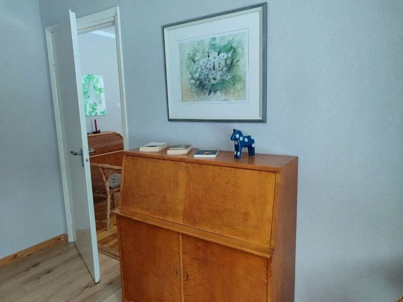 Ein Wohnzimmer mit einer Holz-Kommode, auf der ein blauer Holzzug steht, und mehreren Büchern. An der Wand hängt ein aquarellartiges Gemälde eines Blumenstraußes, darüber eine weiße Rahmen.