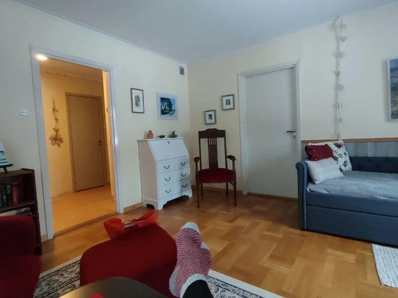 Gemütliches Wohnzimmer mit Holzboden, rotem Sessel, blauer Couch mit Kissen, weißen Wänden und mehreren Bildern an der Wand, Tür zur Diele, kleiner weißer Schrank und einem dunklen Holzstuhl.