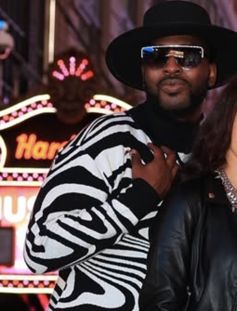 A man wearing a black hat, large sunglasses, a zebra-print jacket, and a black neck scarf standing next to a woman in a black leather jacket. Neon lights and a casino sign are in the background.