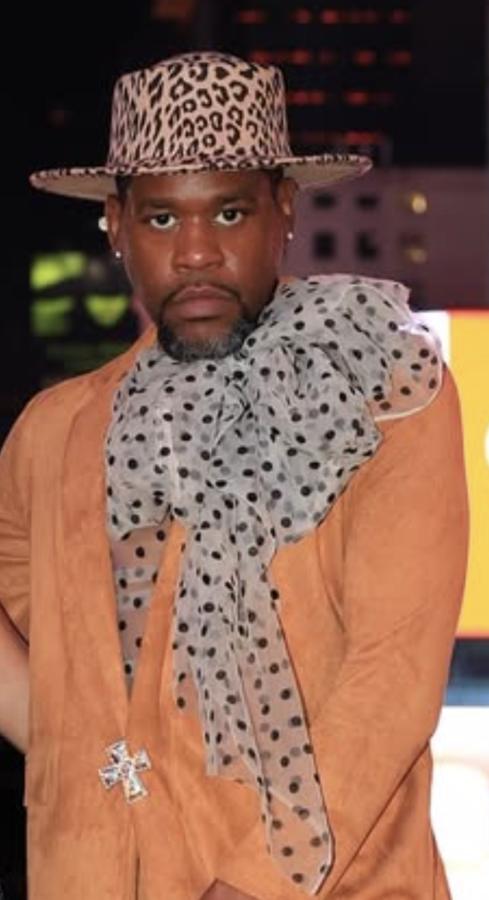 A man wearing a leopard print hat and a polka dot scarf, with a brown jacket, standing in an indoor setting.