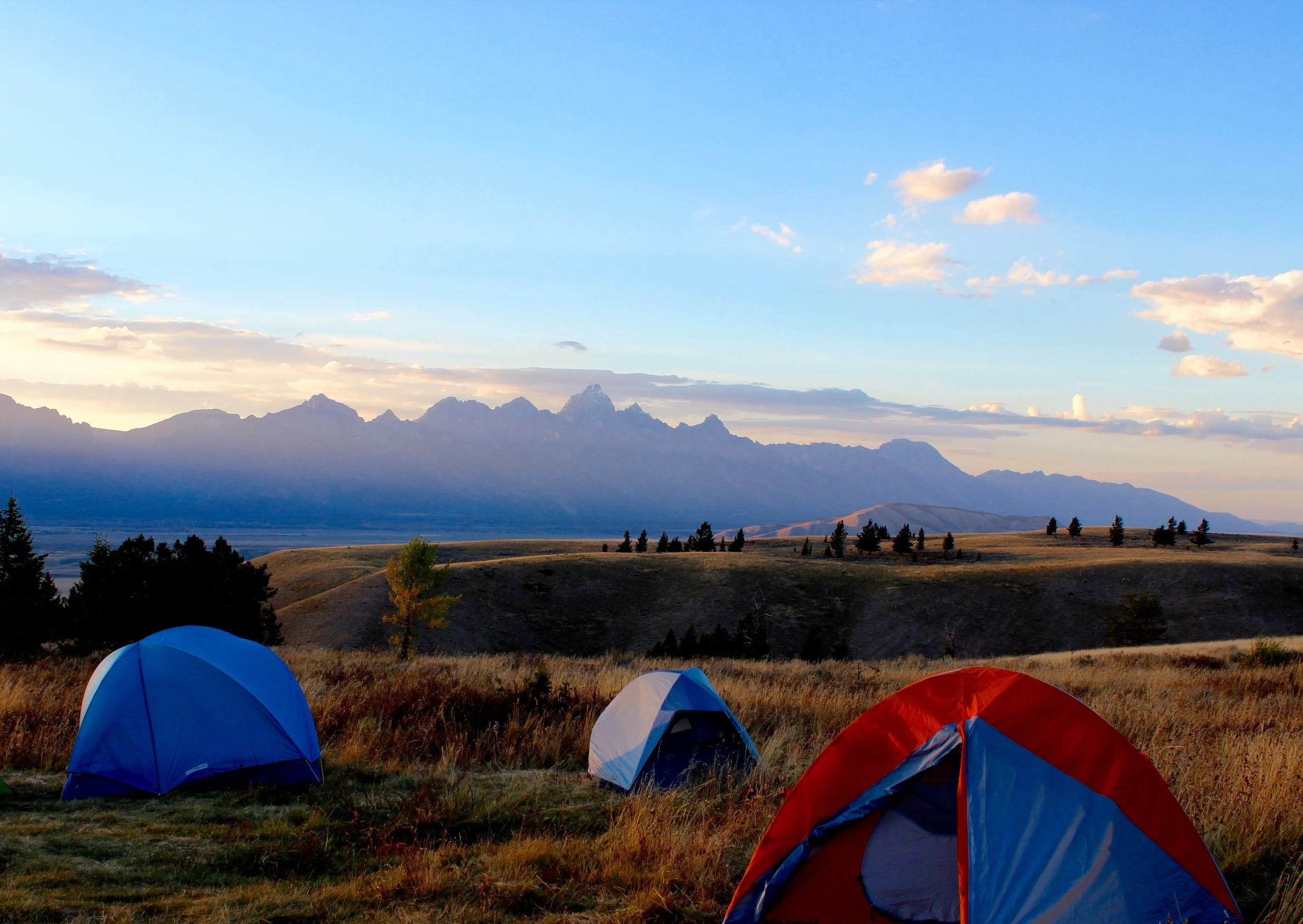 Three colorful tents set up on a grassy hill with a mountain range, trees, and a partly cloudy sky in the background during sunset.