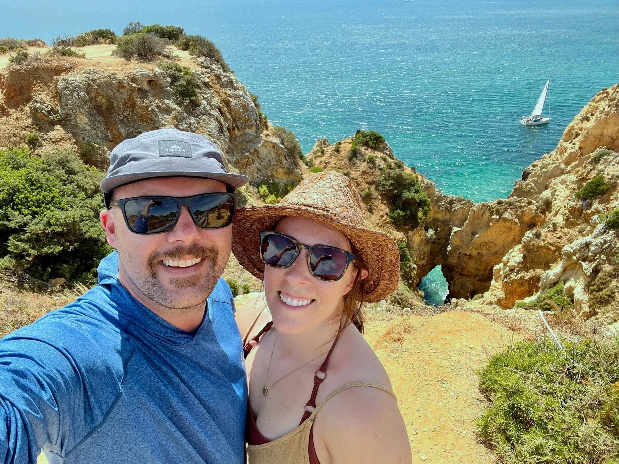 A smiling man and woman taking a selfie on a cliff overlooking the ocean with a sailboat, rocky formations, and a natural arch in the background.