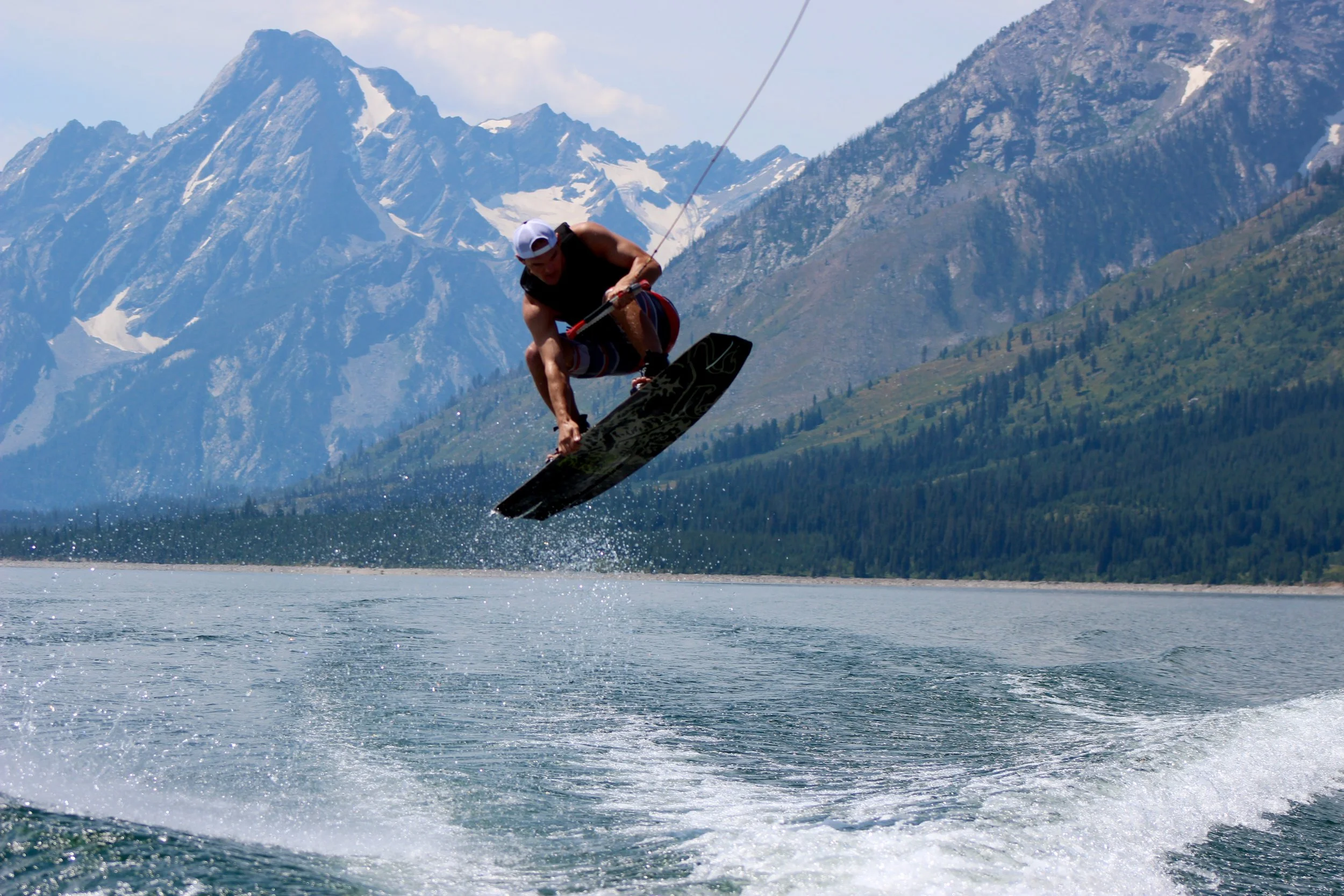 Person wakeboarding on a lake with snow-capped mountains and forested hills in the background.