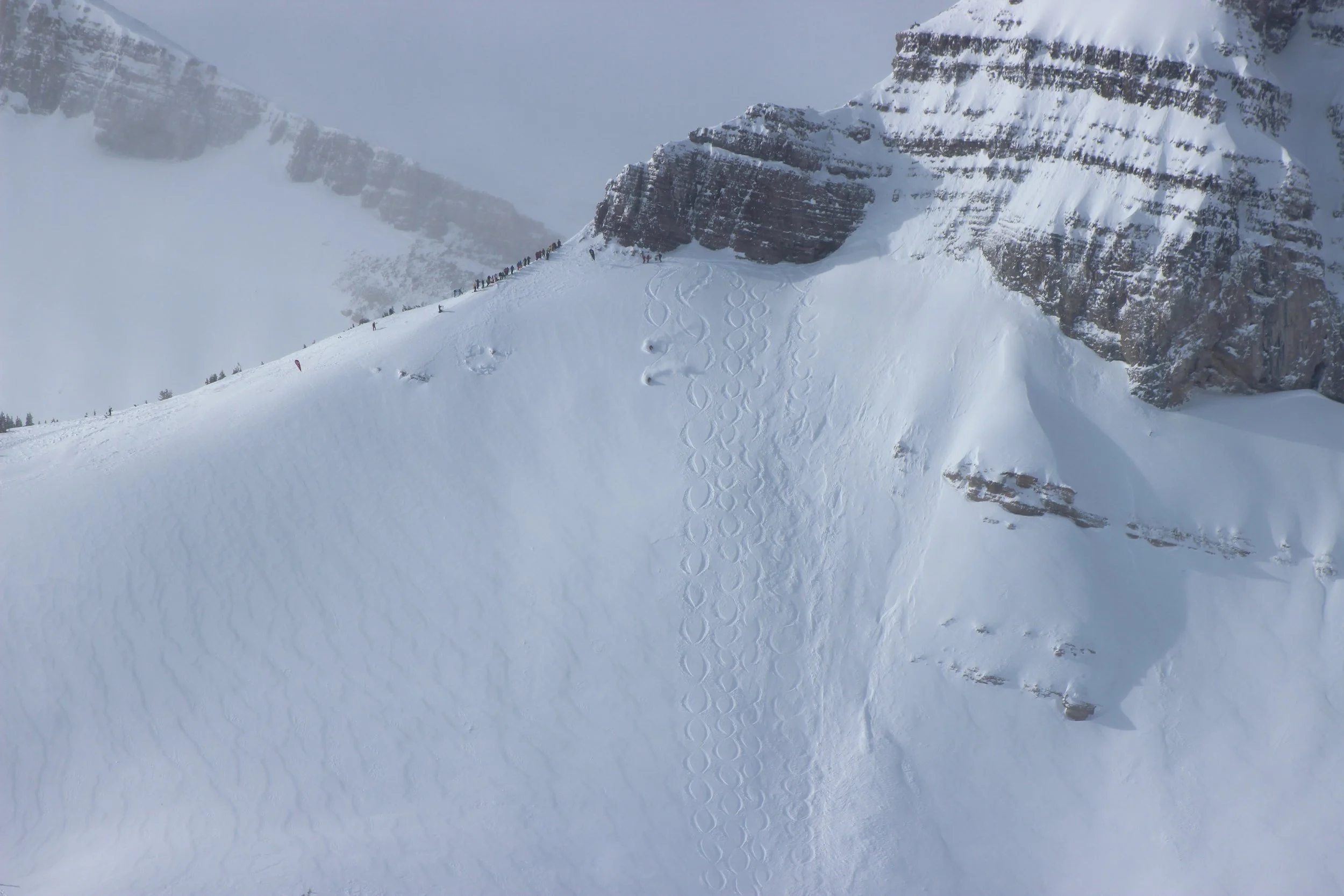 A large snow-covered mountain slope with ski or snowboard tracks and a group of people at the top of the slope.