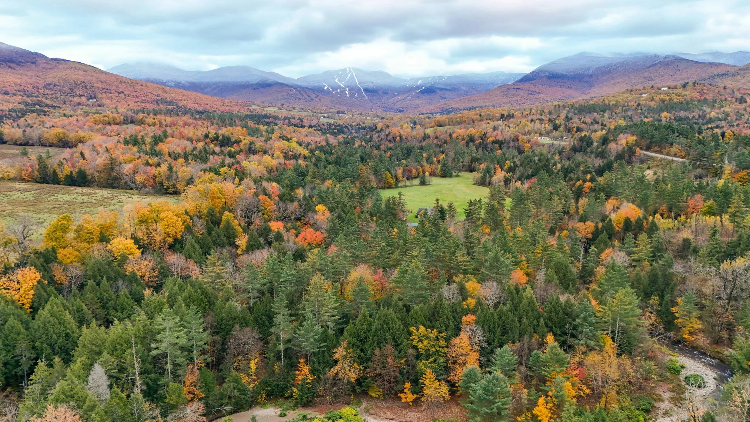 Aerial view of a forested valley during autumn with colorful foliage, mountains in the background, and a partly cloudy sky.
