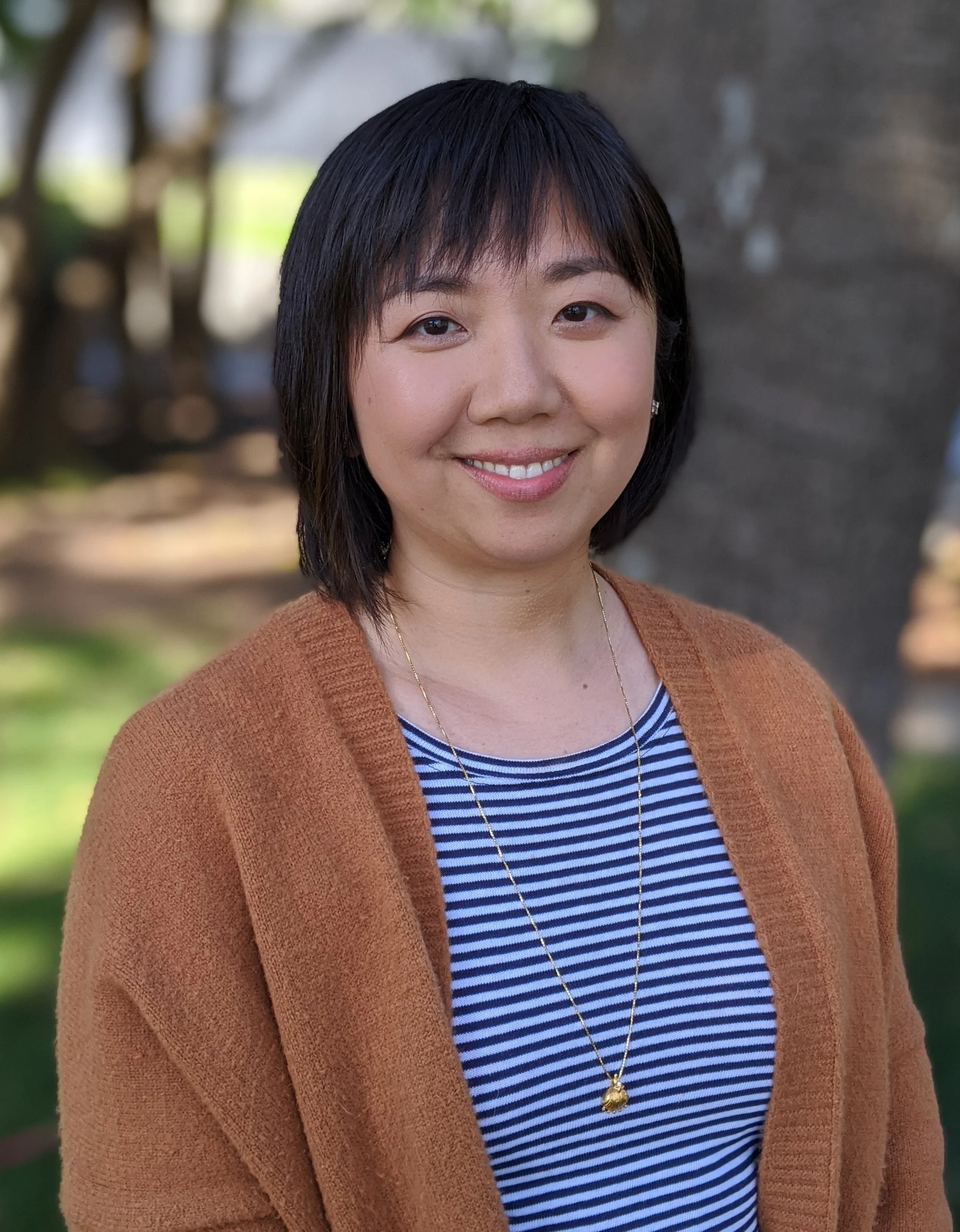 A woman with short black hair smiling outdoors, wearing a striped blue and white shirt, a brown cardigan, and a gold necklace.