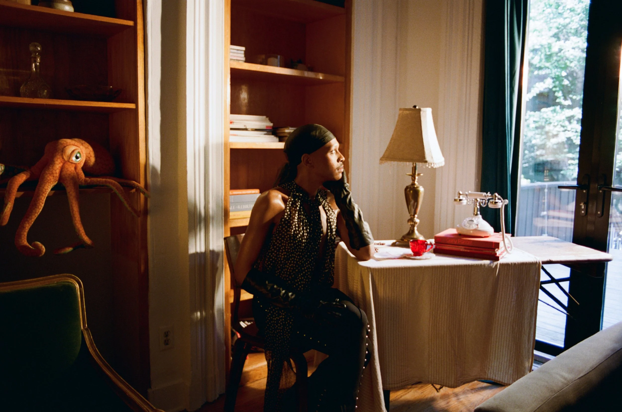 A woman with braided hair sitting at a dining table in a sunlit room, with a lamp, a pile of books, and a window with blue curtains in the background.