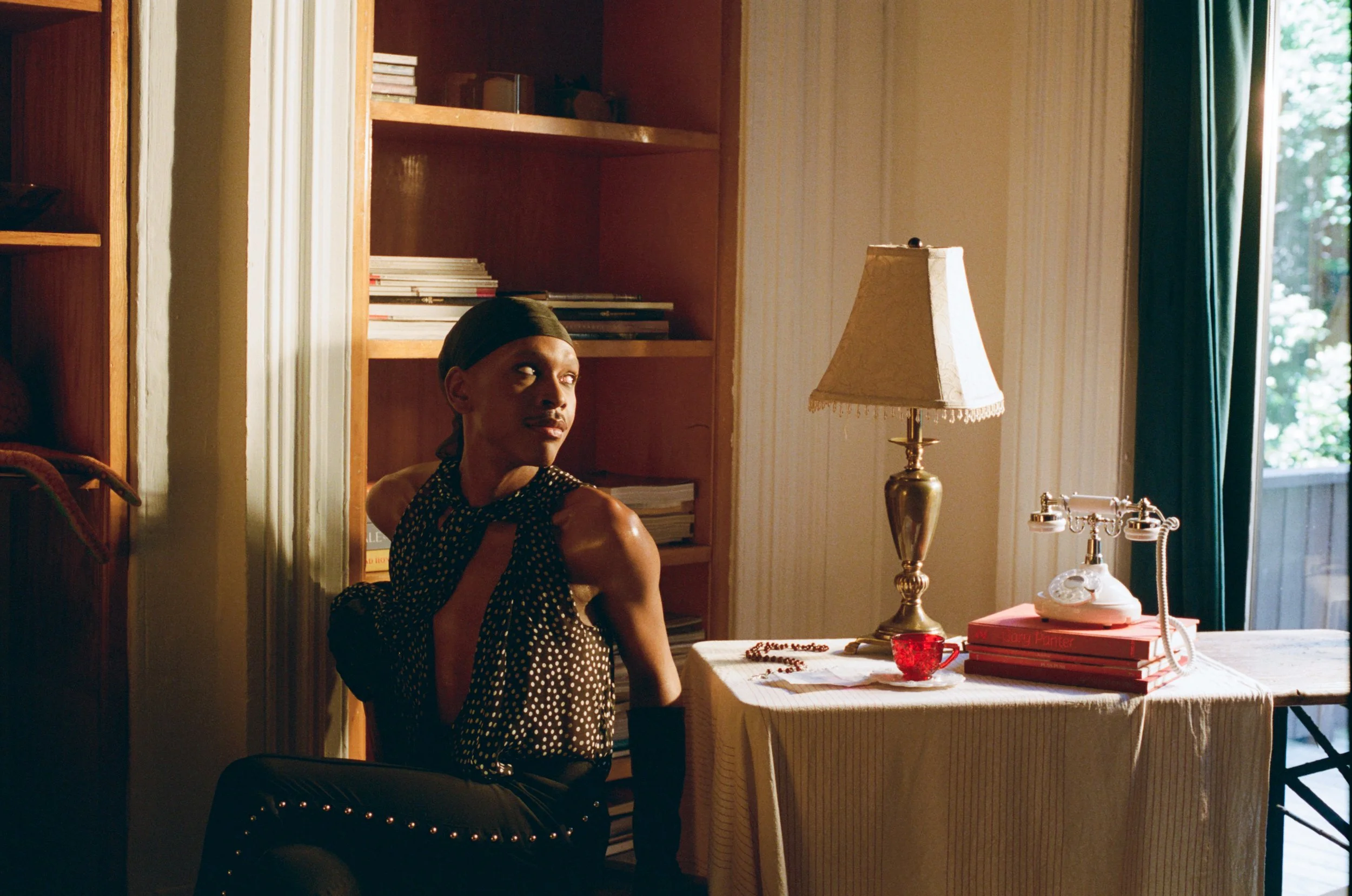 A woman with dark hair wearing a polka dot dress and a headscarf sitting in a sunlit room next to a table with a vintage telephone, a stack of books, a lamp, and a jewelry necklace.