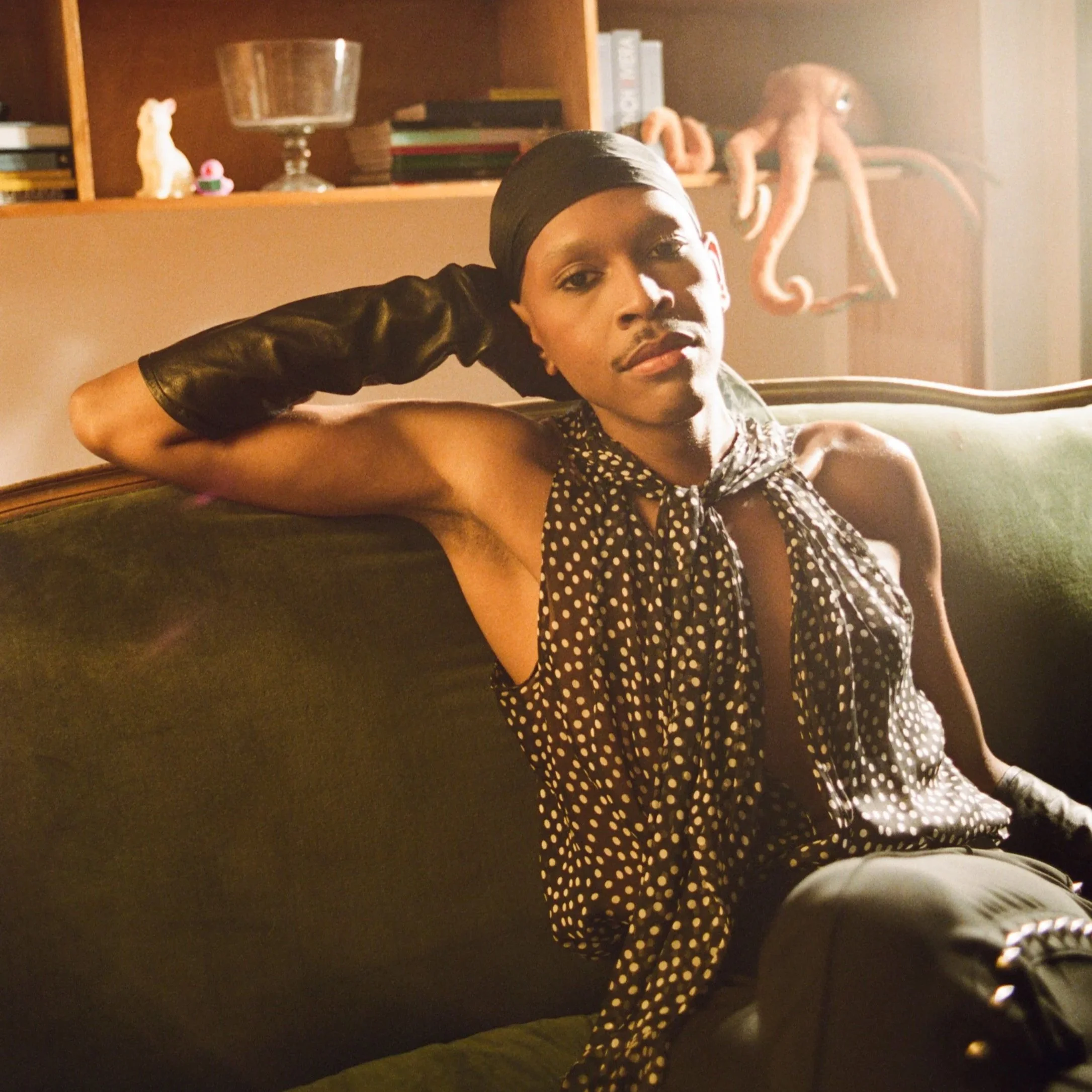 A person sitting on a couch with their arm resting on the back, wearing a polka-dot dress and a black headscarf, looking at the camera with a relaxed expression. In the background, there is a bookshelf with decorative items, magazines, and a large pink octopus sculpture.