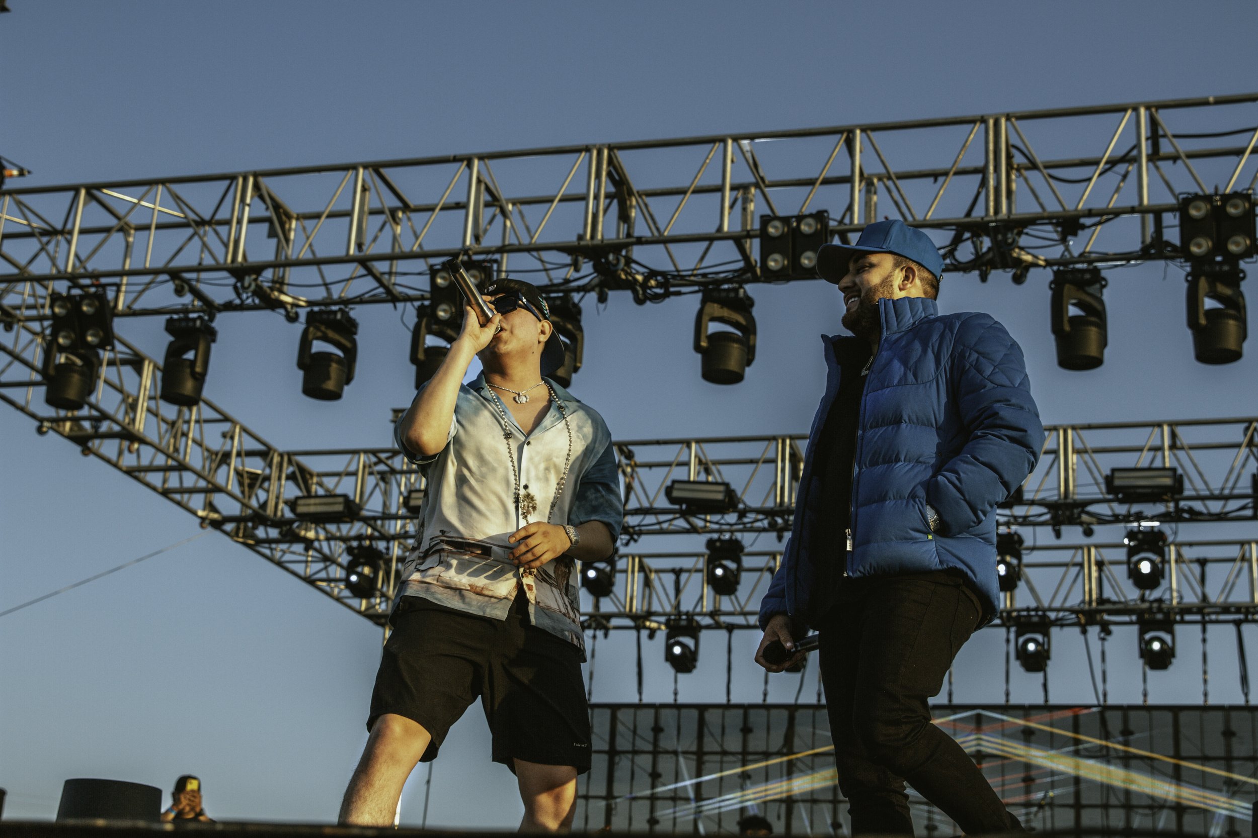 Two men standing on an outdoor stage with lighting rigs above, one singing into a microphone and the other listening, during sunset.