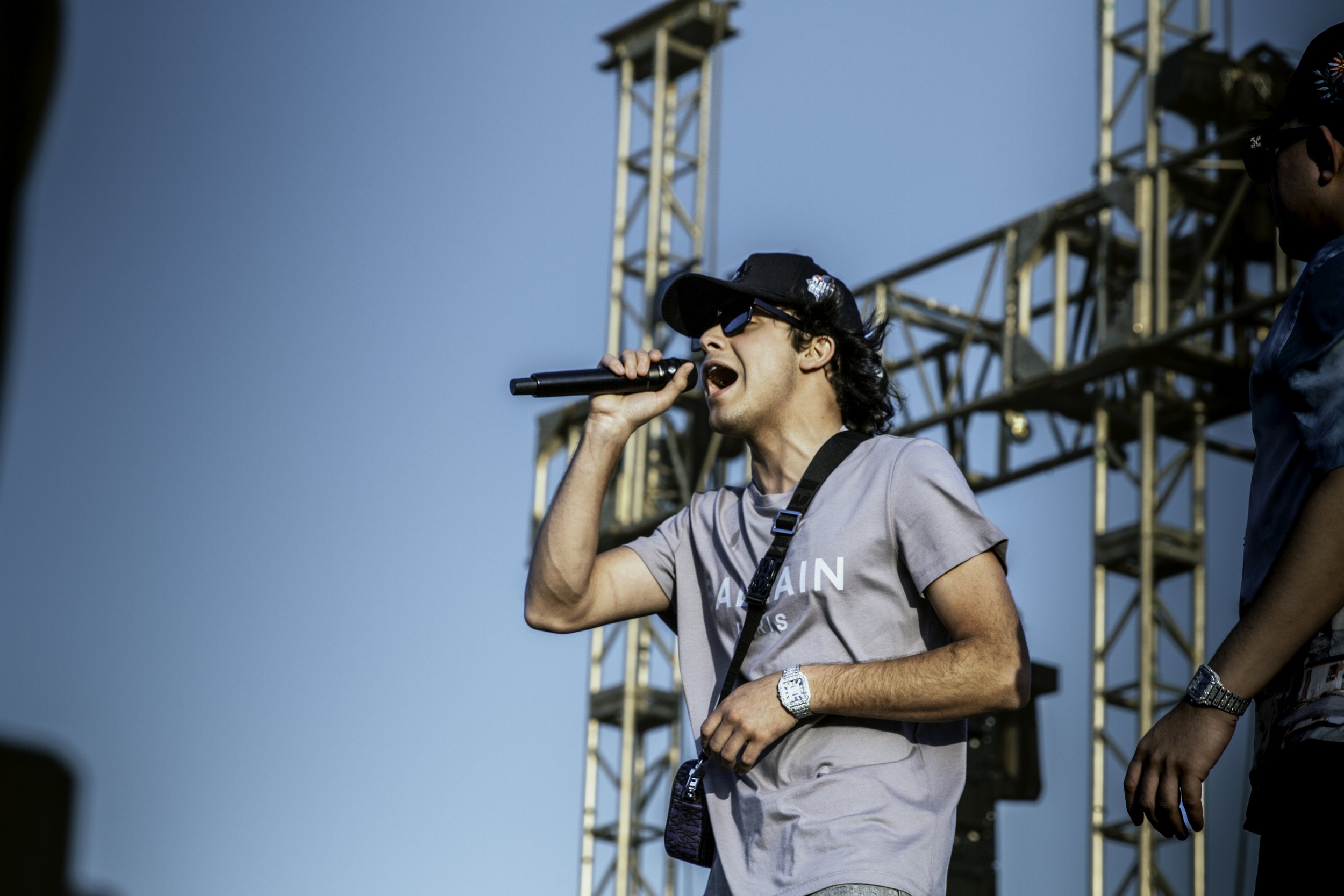 A man wearing sunglasses and a gray T-shirt singing into a microphone at an outdoor event during the daytime with a blue sky and stage scaffolding in the background.