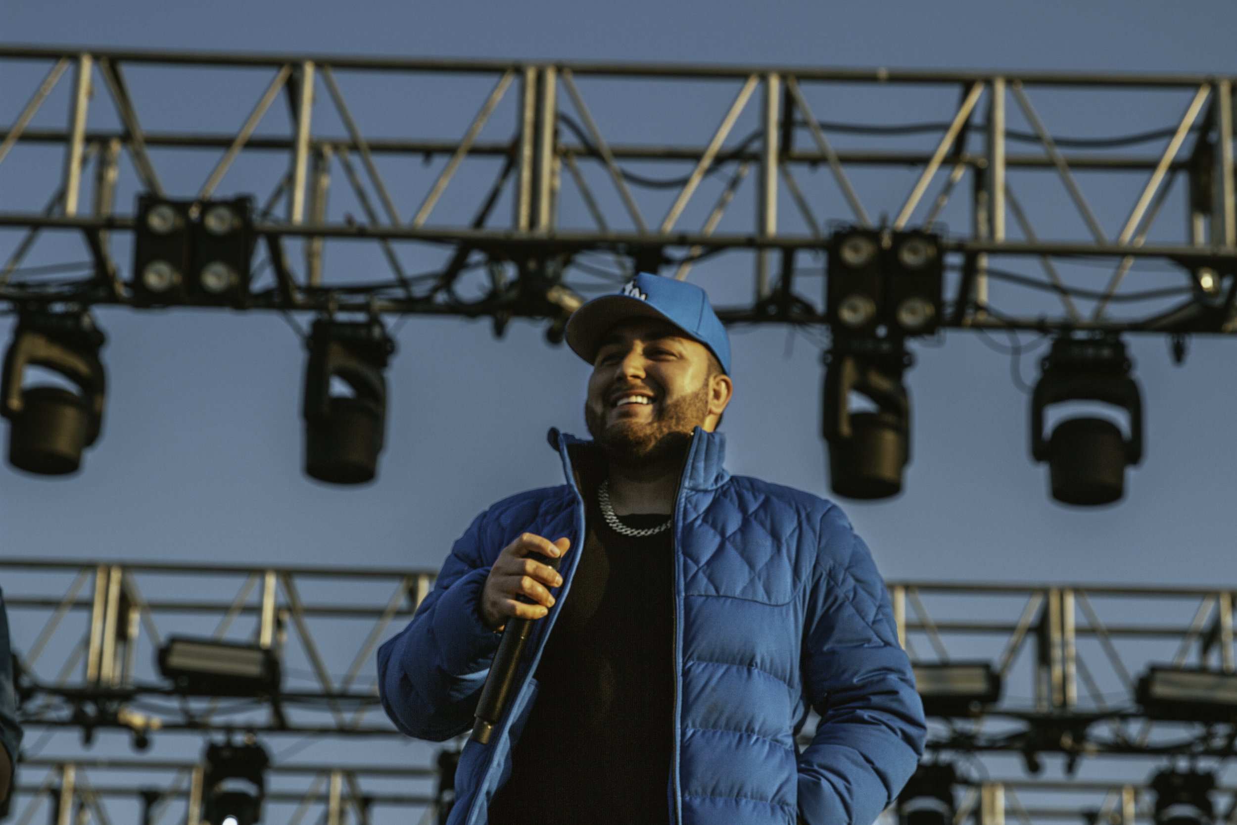 A man wearing a blue jacket, cap, and the chain holding a microphone, standing under a stage with lighting equipment during daytime.
