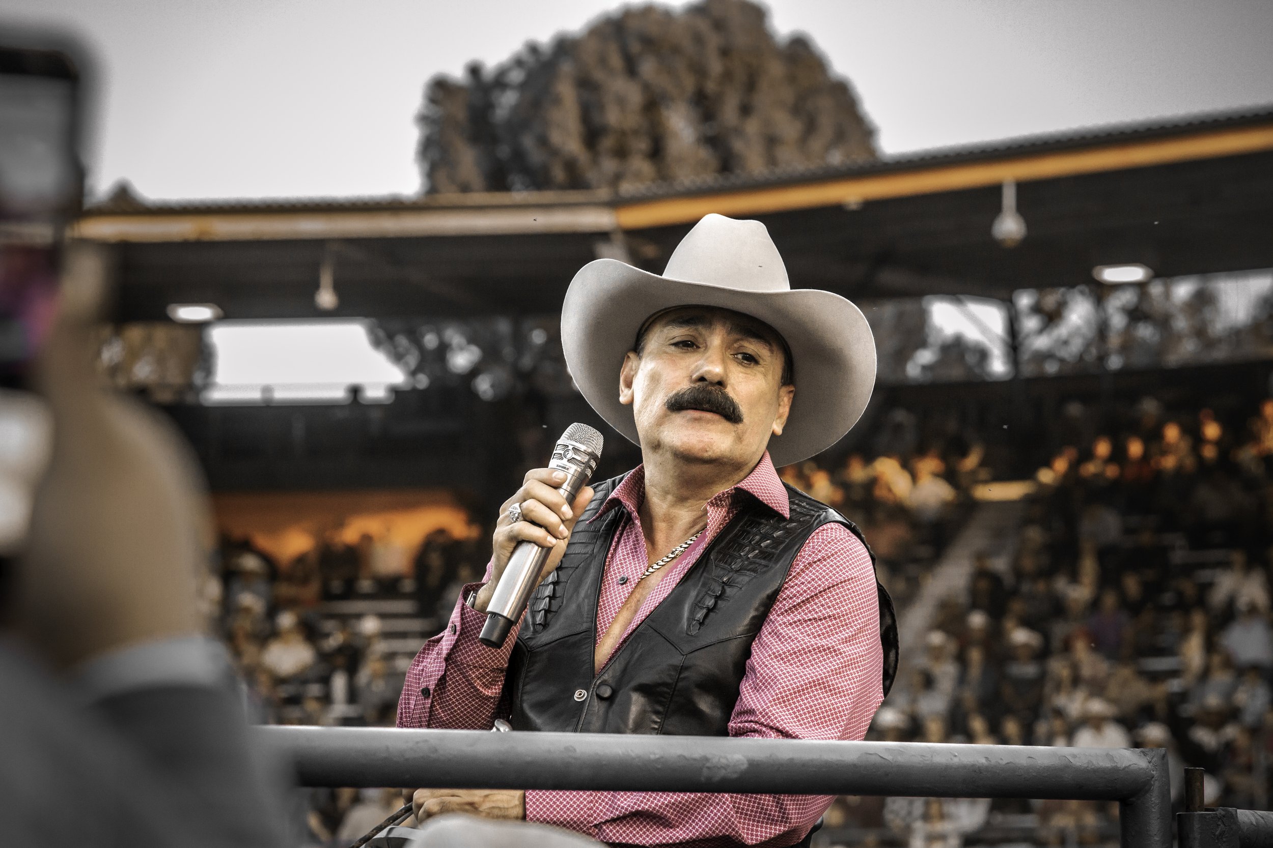 A man wearing a cowboy hat and a leather vest holding a microphone, speaking at an outdoor event with a crowd in the background.