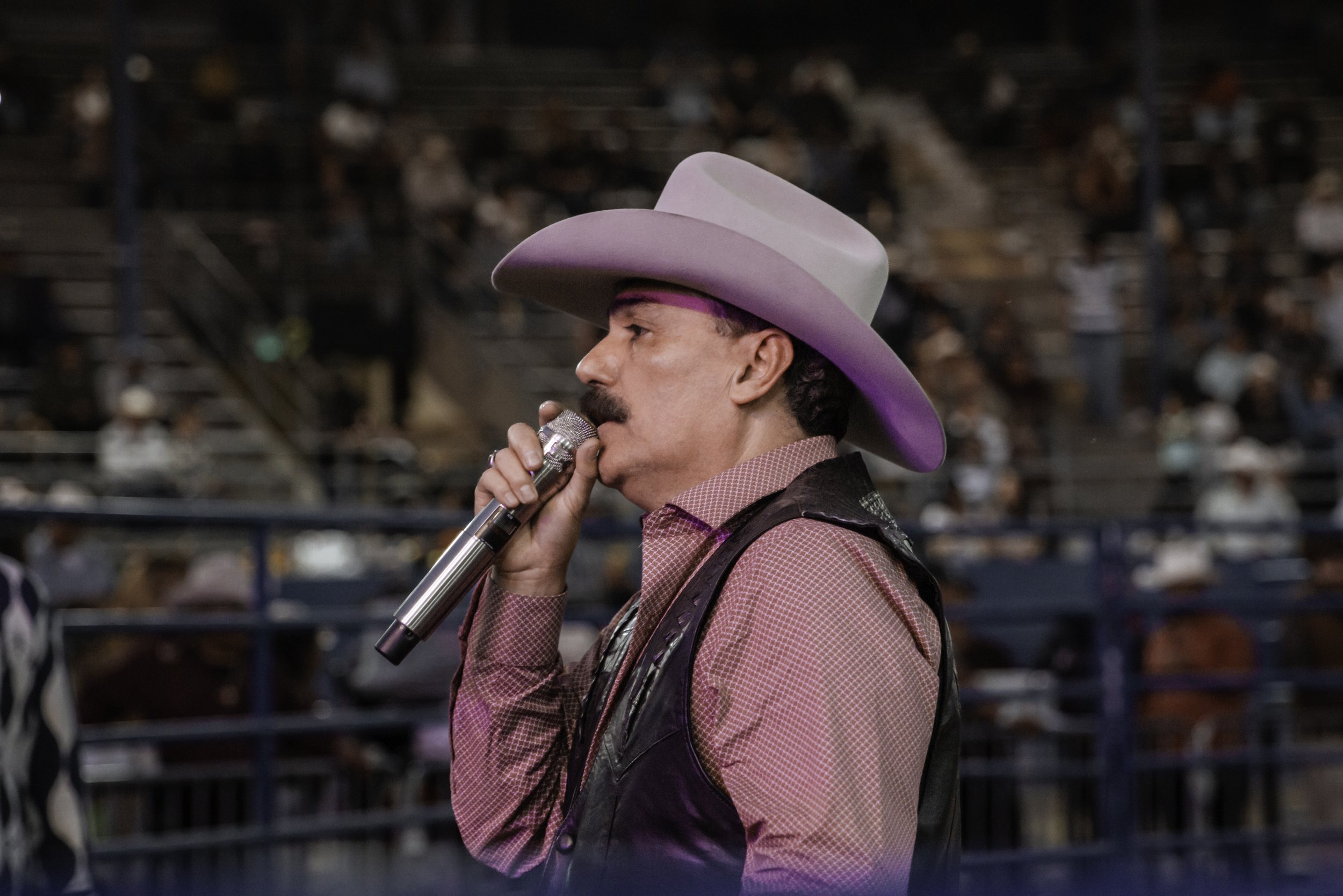 A man with a mustache wearing a cowboy hat and a pink patterned shirt, holding a microphone and speaking at an event with a crowd in the background.