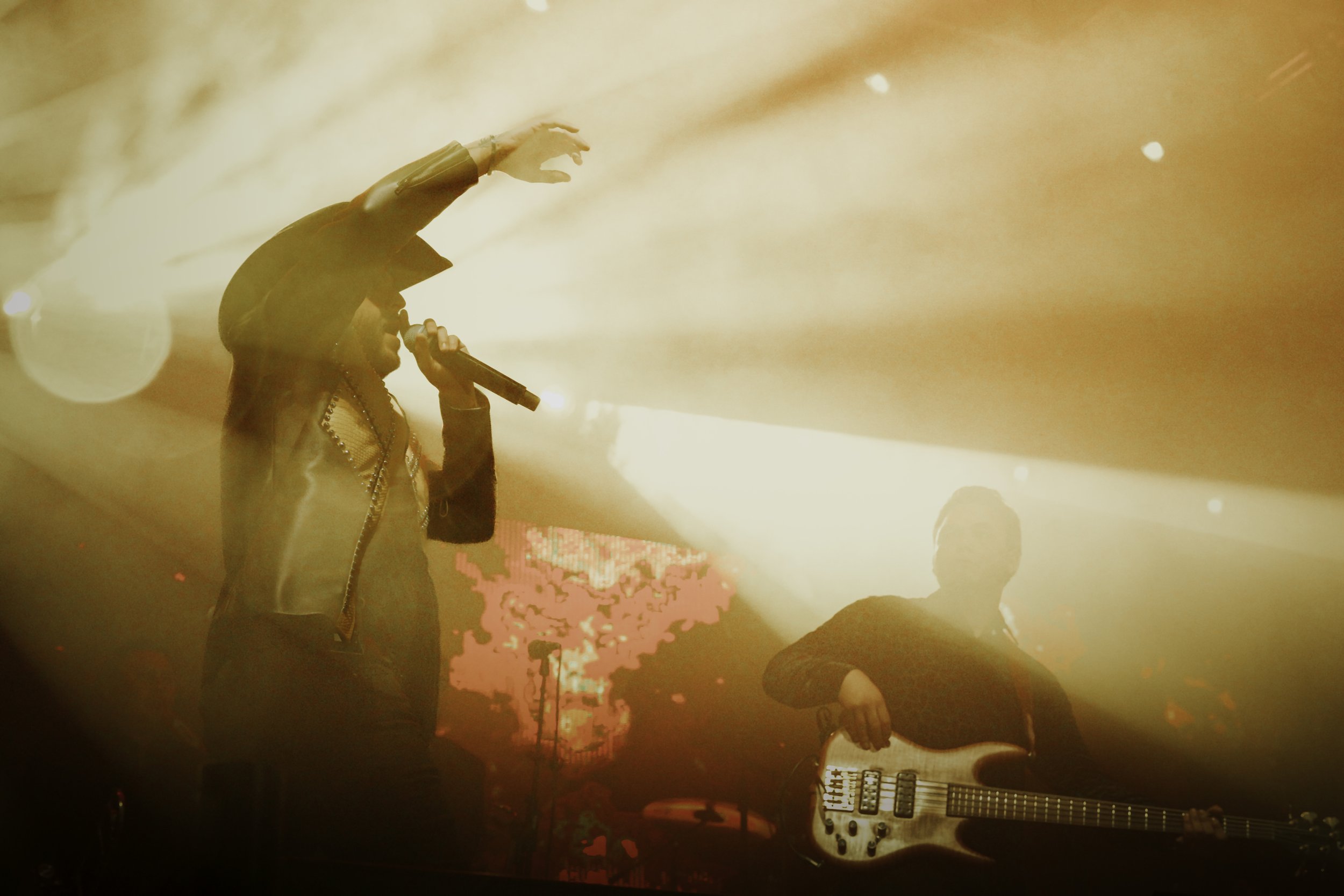Musicians performing on stage, with one singing into a microphone and another playing a bass guitar, under bright, warm stage lighting.