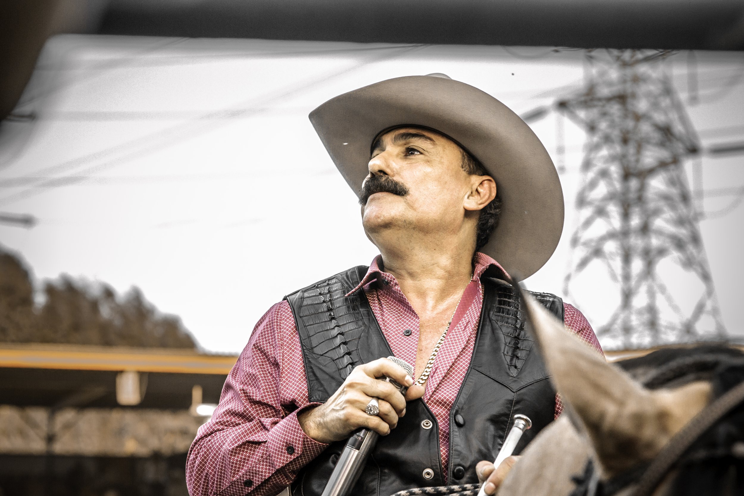 A man wearing a cowboy hat, red checkered shirt, and black leather vest standing outdoors. He has a mustache and is holding a microphone with a large power line tower visible in the background.
