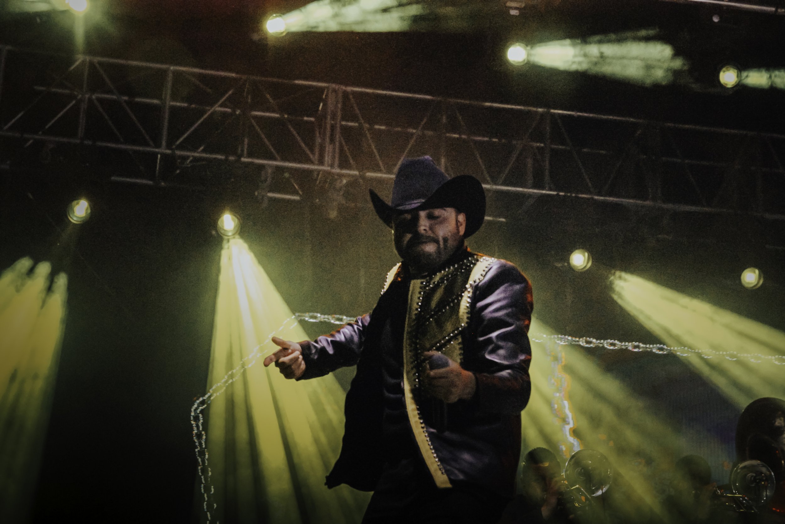 Performing artist on stage wearing a black hat and cowboy jacket with gold accents, with yellow stage lights illuminating the scene.