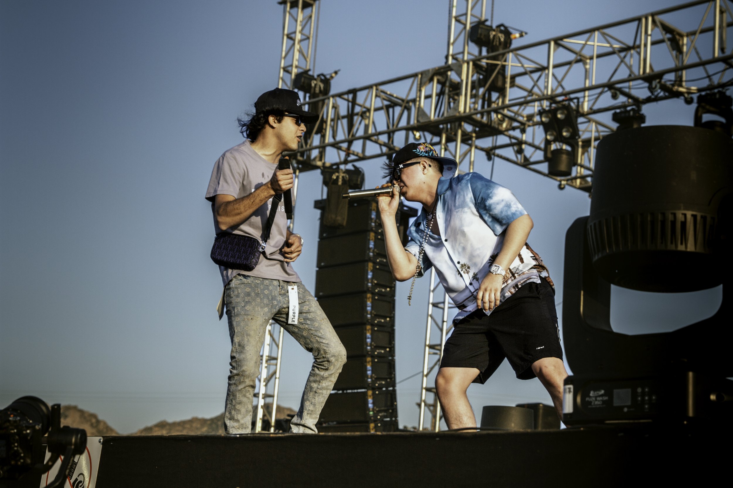 Two men performing on an outdoor stage, one holding a microphone and the other leaning in to sing or speak, with stage lighting and equipment behind them.