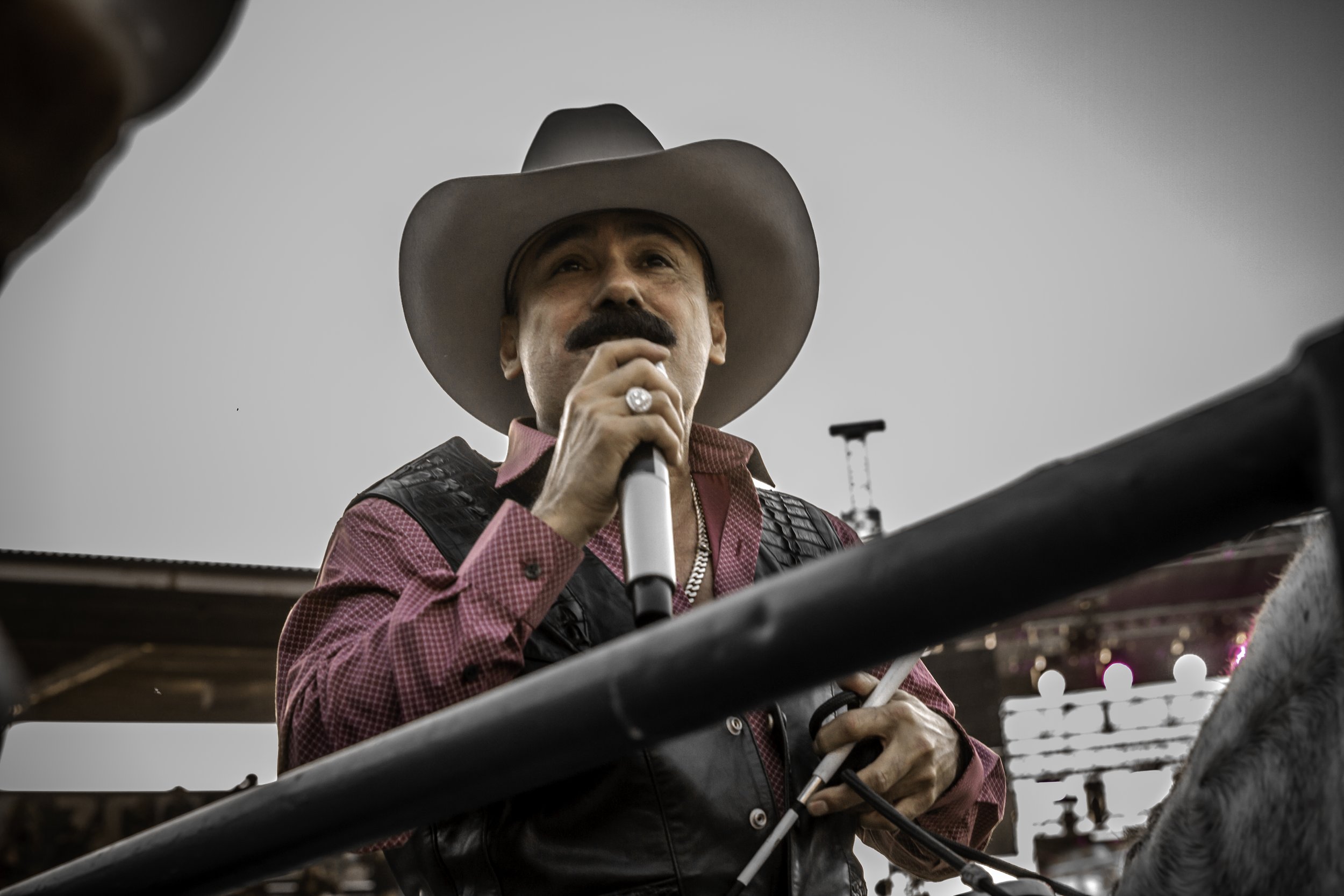 A man wearing a cowboy hat, red shirt, and leather vest speaking into a microphone at an outdoor event, with a cloudy sky in the background.