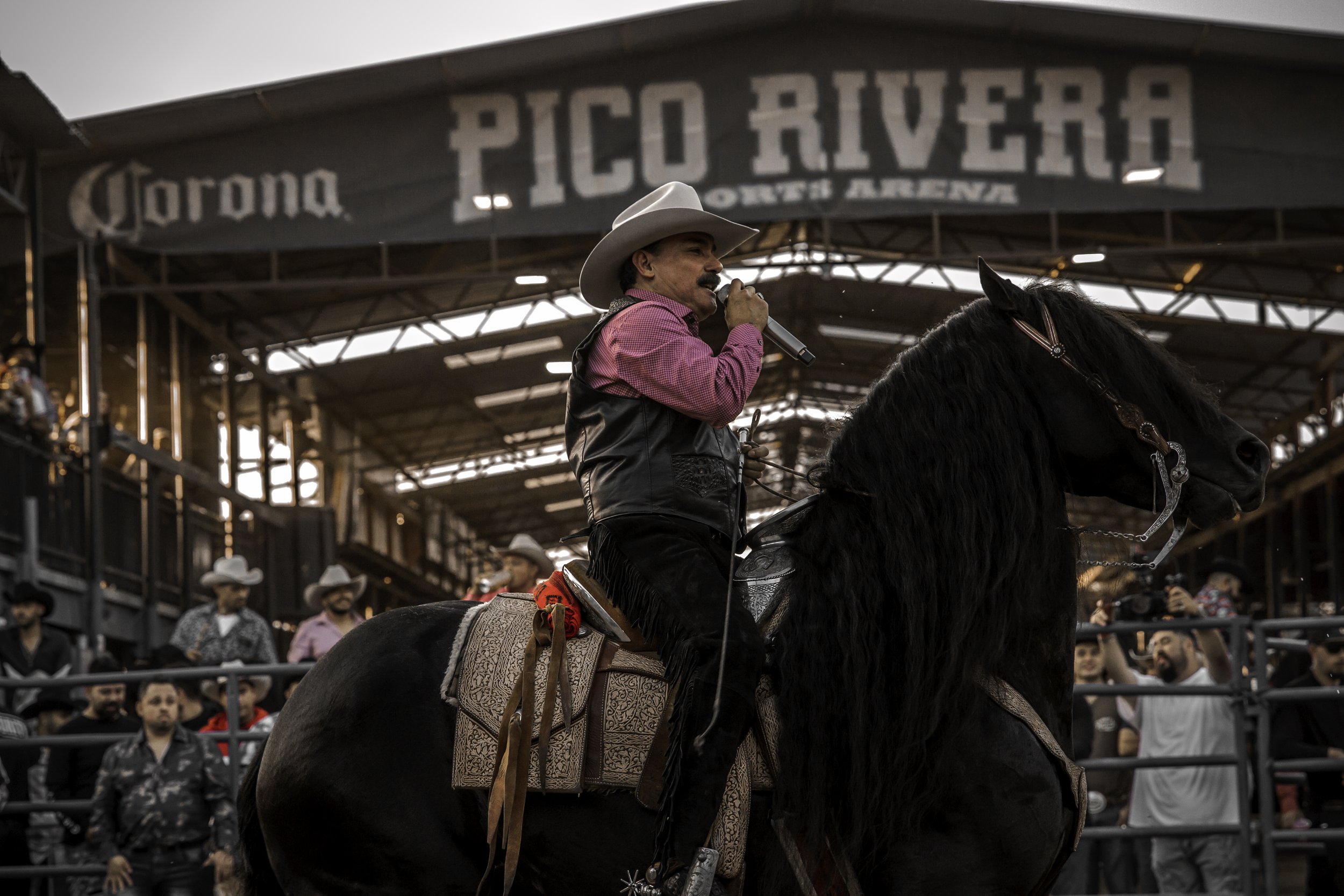 A cowboy dressed in a pink shirt and black vest sits on a black horse, speaking into a microphone at an arena event. The background shows a large sign reading 'Pico Rivera' and a crowd of spectators, some wearing cowboy hats.