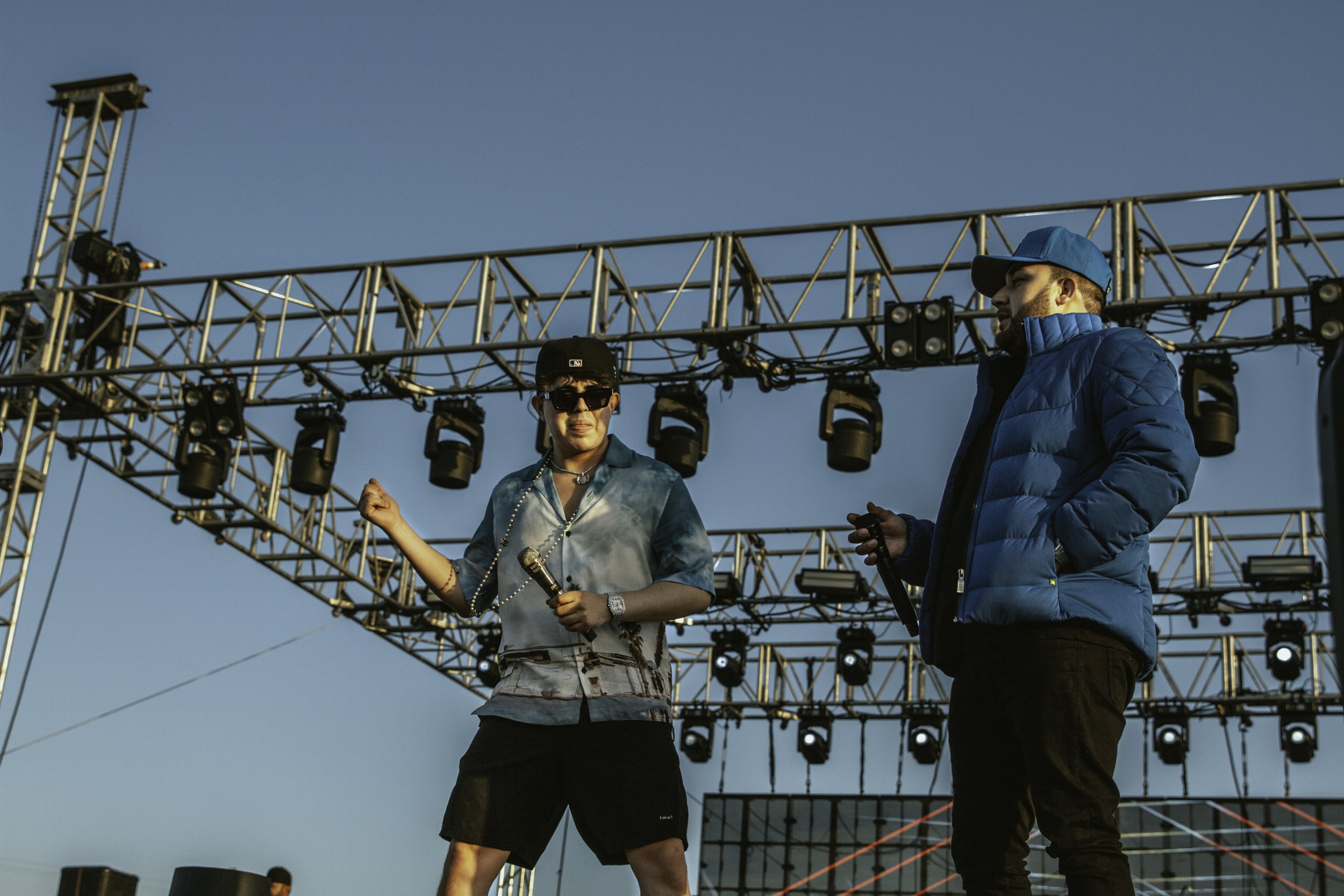 Two male performers on an outdoor stage with metal truss structure, one holding a microphone and wearing sunglasses, the other holding a microphone and wearing a blue puffer jacket and cap, during a concert in the daytime.
