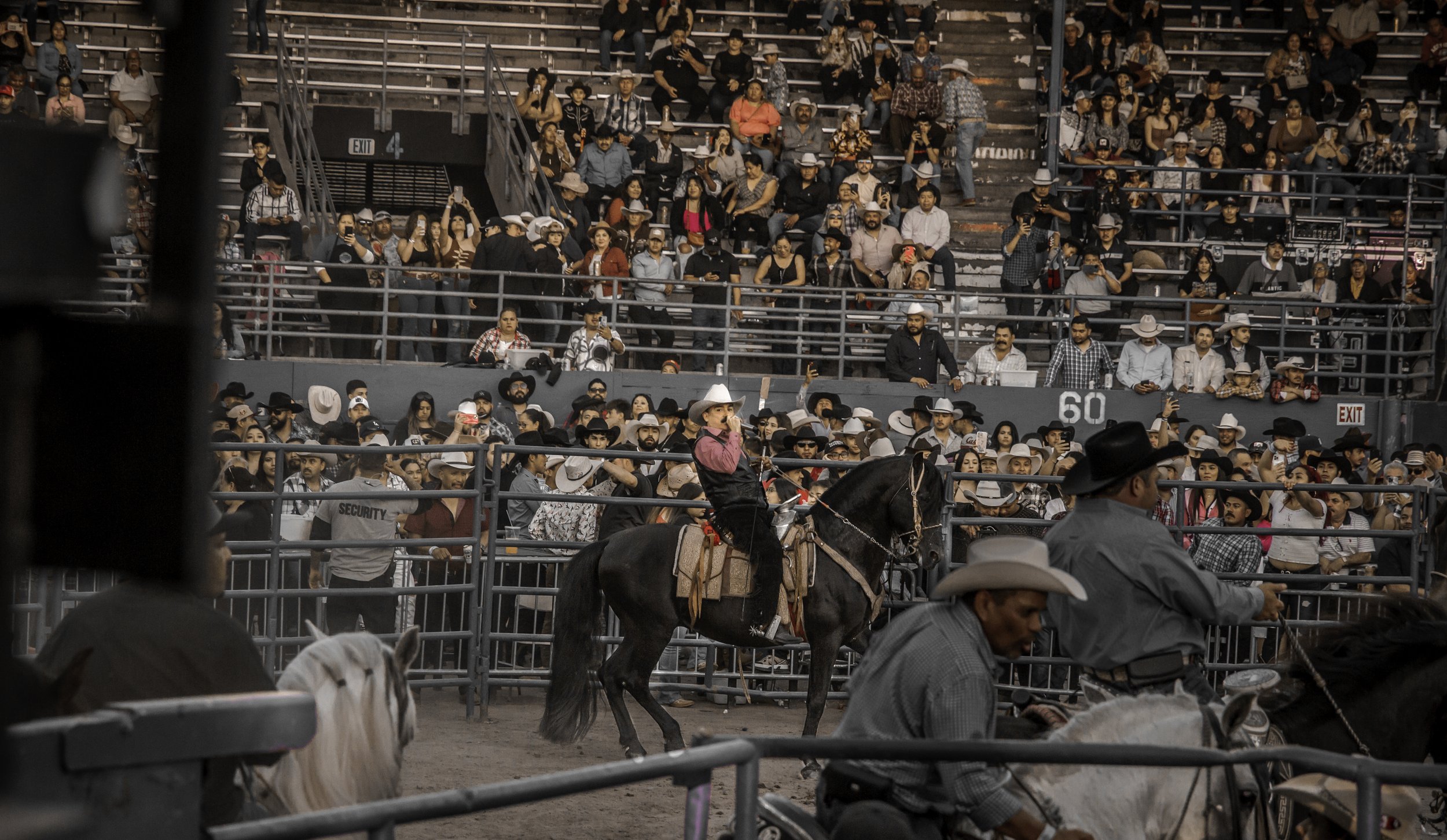 A rodeo event with a rodeo clown on a black horse, surrounded by cowboys on horseback and a large audience in the stands.