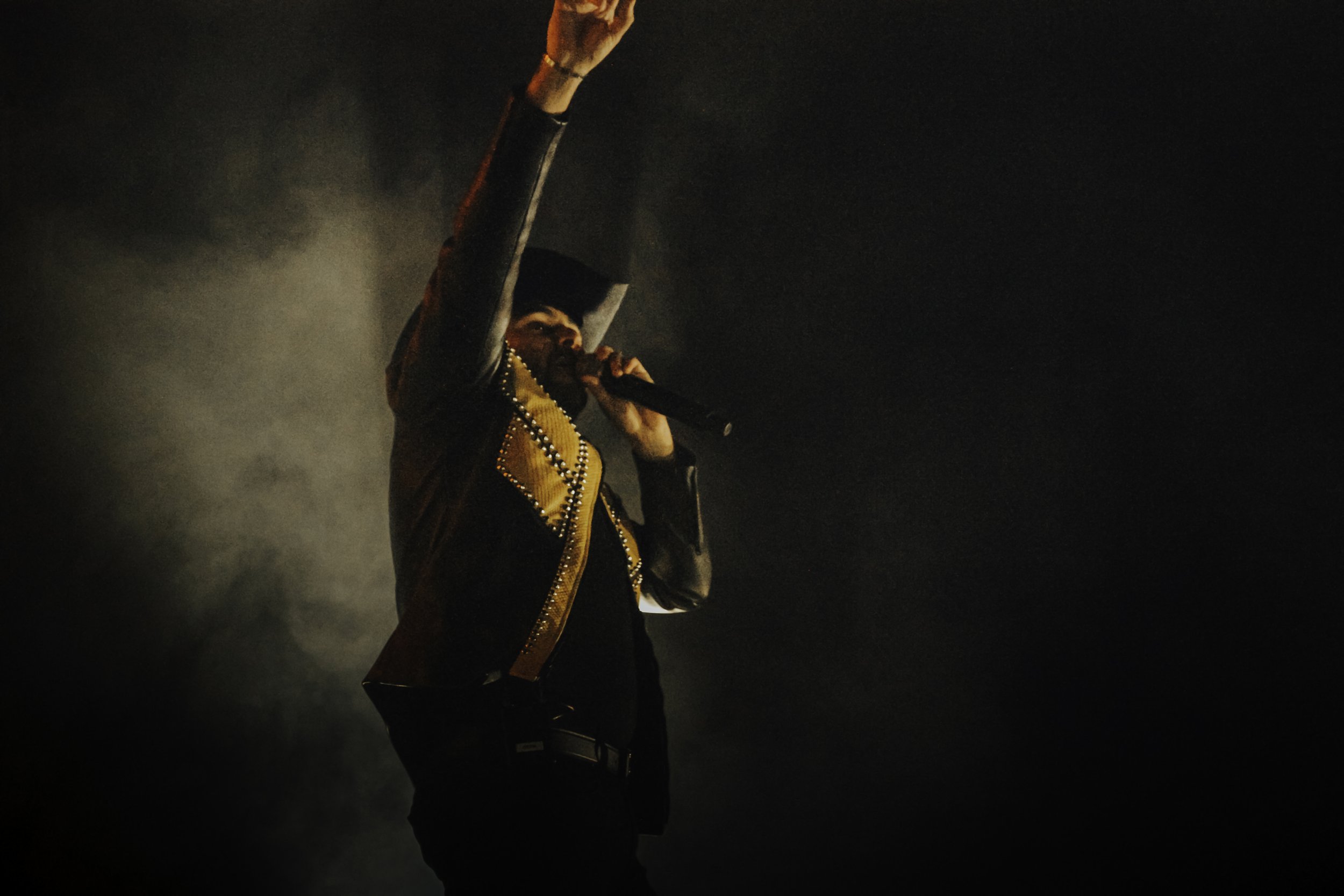 Performer wearing a traditional Mexican charro jacket, singing or speaking into a microphone with one arm raised, on a dark stage with smoke.