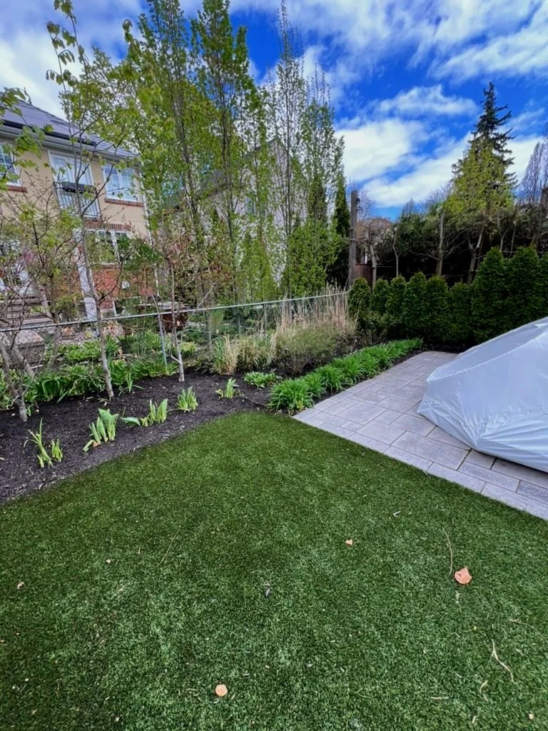 A backyard garden with a vibrant green lawn, flower bed, trees, and a partly cloudy sky.