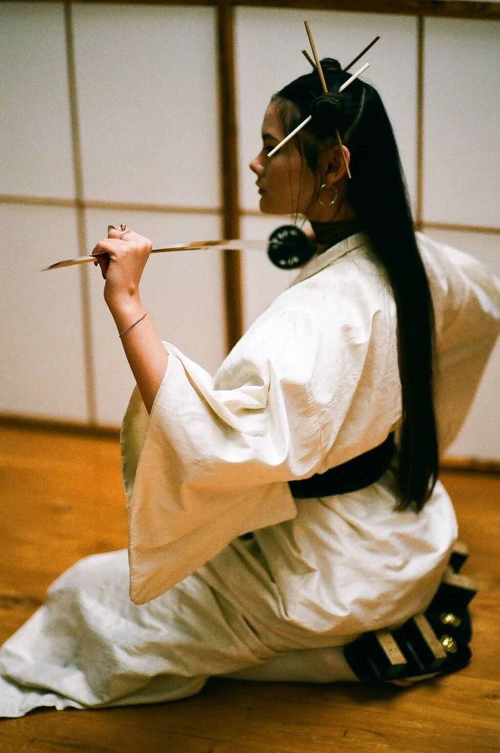 A photo shot of a young Asian female model. The model wears a cream kimono and traditional Japanese attire. Her hair is down, but up at the front in buns with sticks penetrating through them. She holds a black samurai sword in her hand.