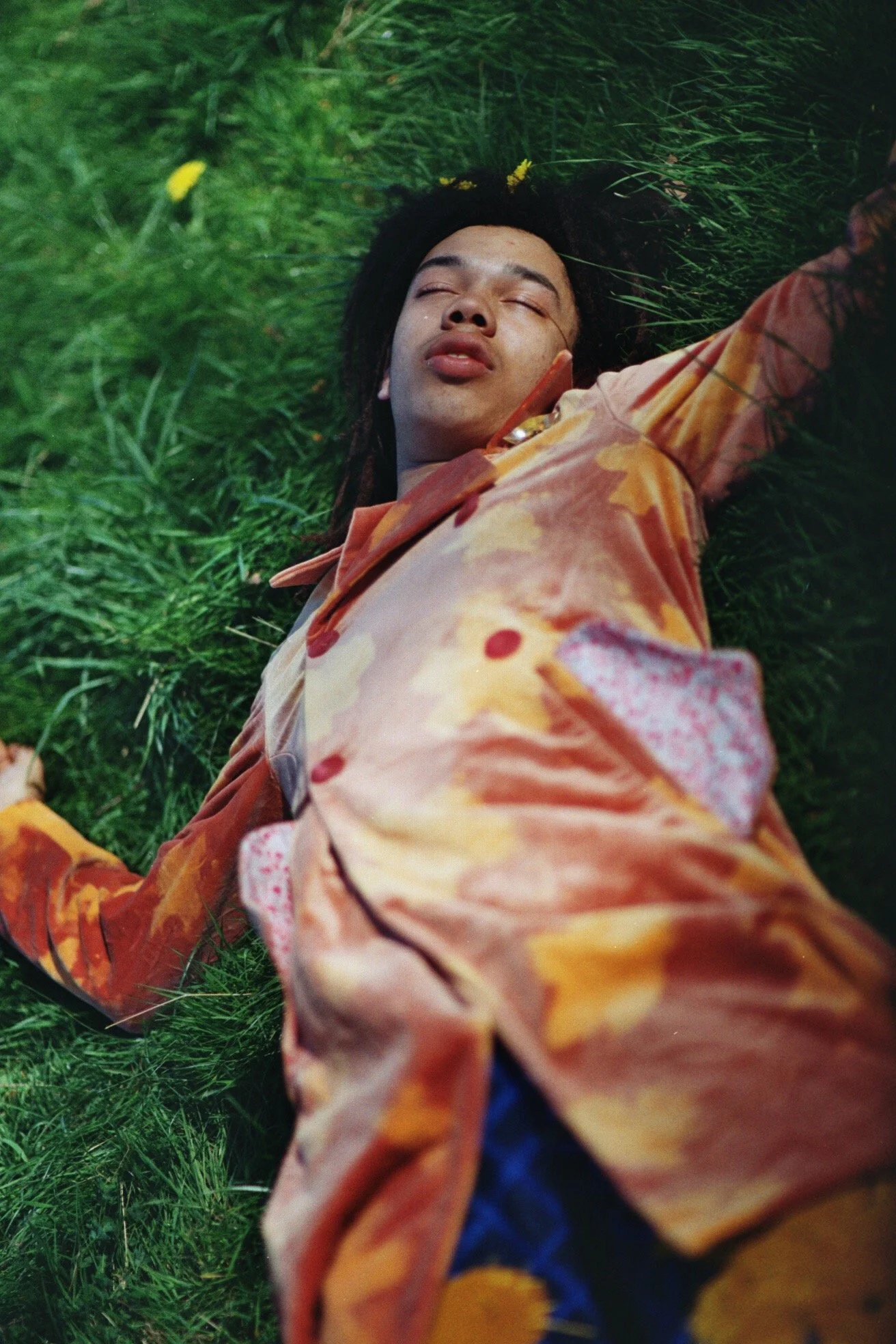 A photo of musician Cosmo Pyke shot from above, laying in a field of grass. He is wearing of velvet peacoat with a floral pattern. His eyes are closed.