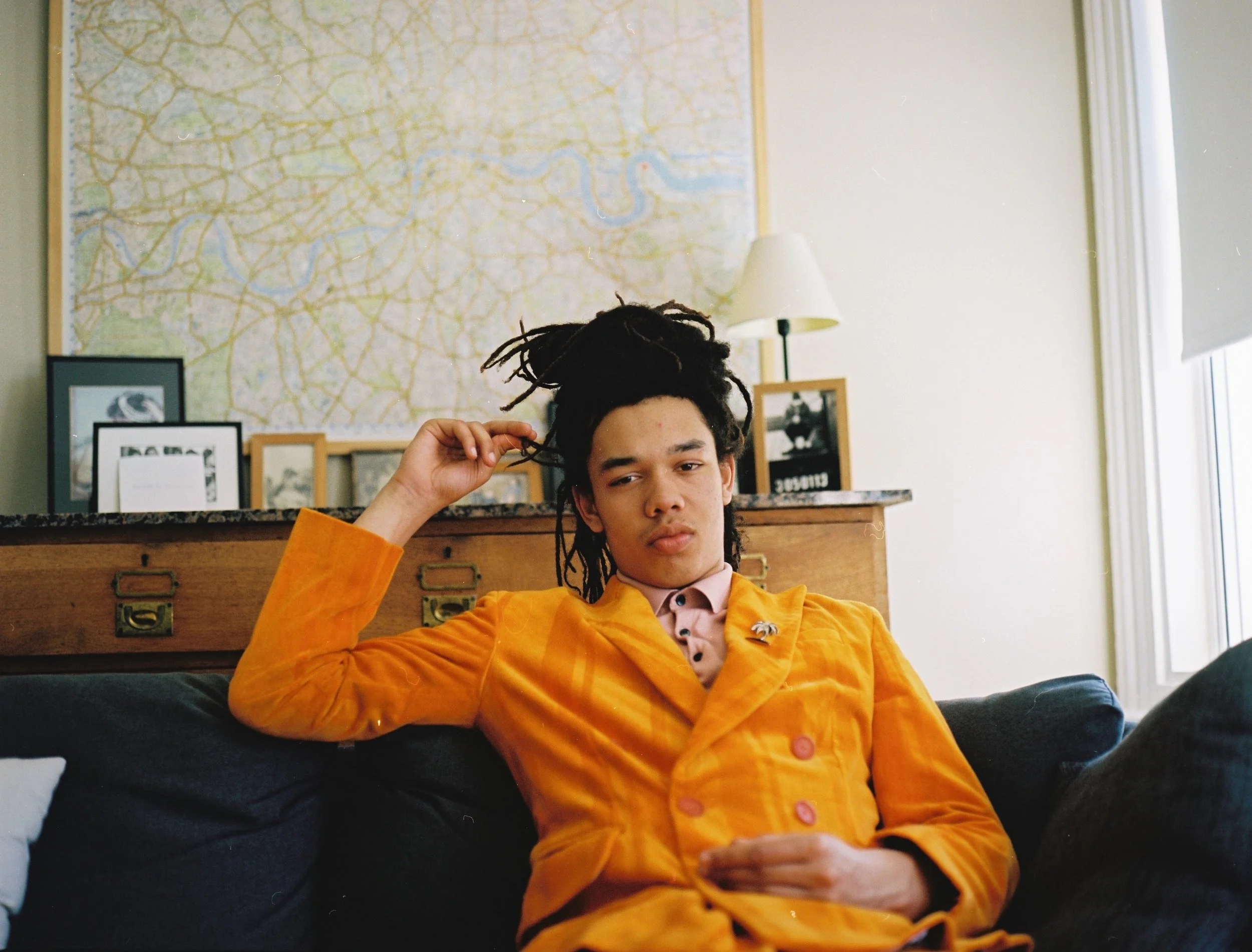 A photo of musician Cosmo Pyke sat in a modern living room. He was wearing an orange velvet suit jacket and pink shirt. He touches one of the locks tied up on his head.