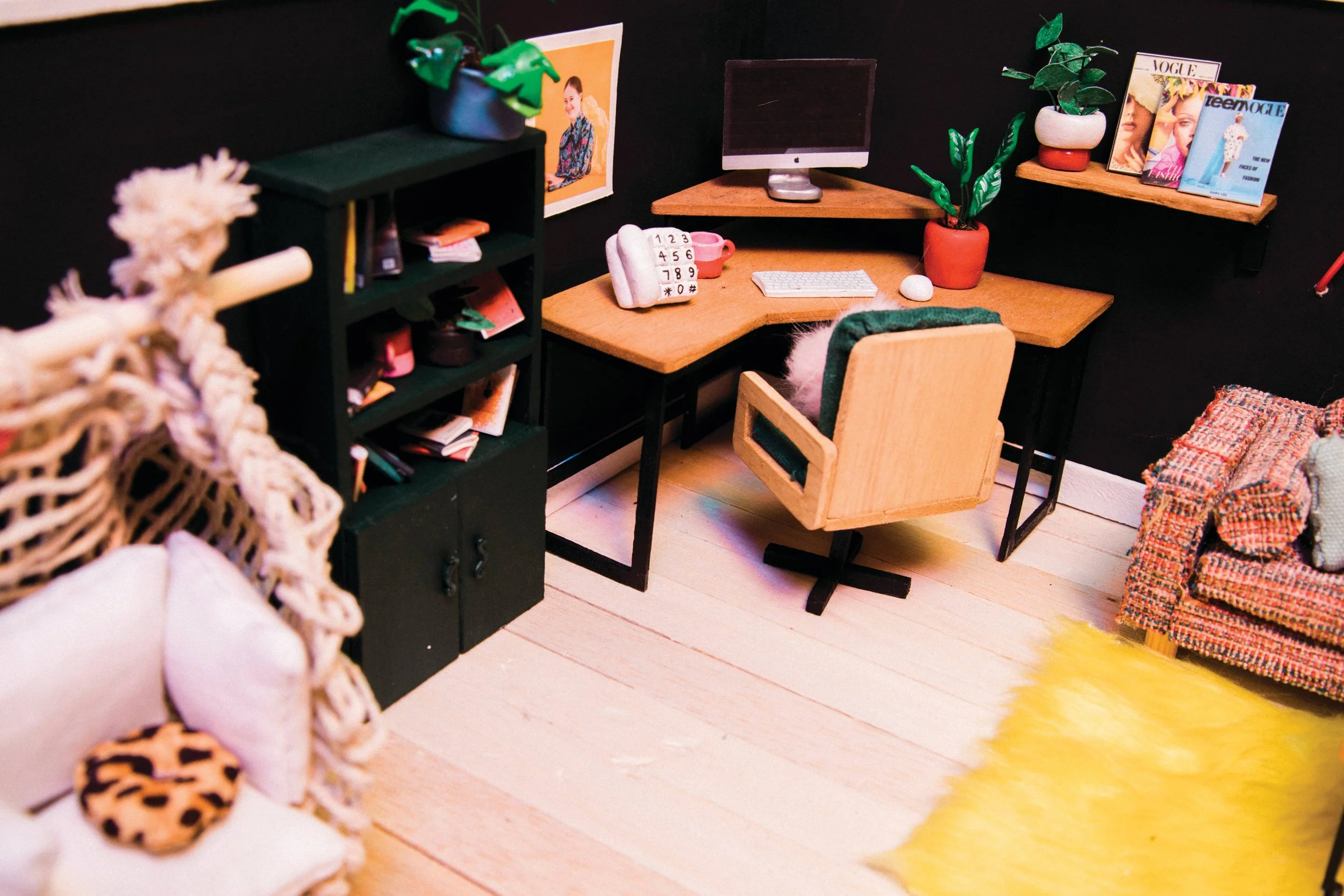 A miniature set of furniture arranged like a home office. There is a wooden desk and brown chair situated in the back right corner of a black room with wood paneled floors. To the left of the frame is a black bookshelf with a green plant resting on t