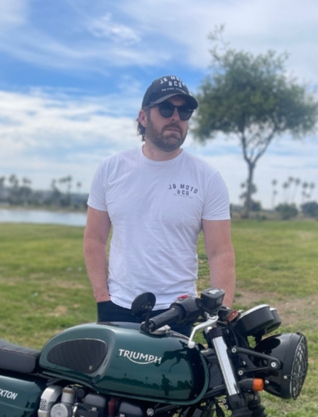Man wearing sunglasses, a black cap, and a white t-shirt standing outdoors next to a Triumph motorcycle with a lake and trees in the background.