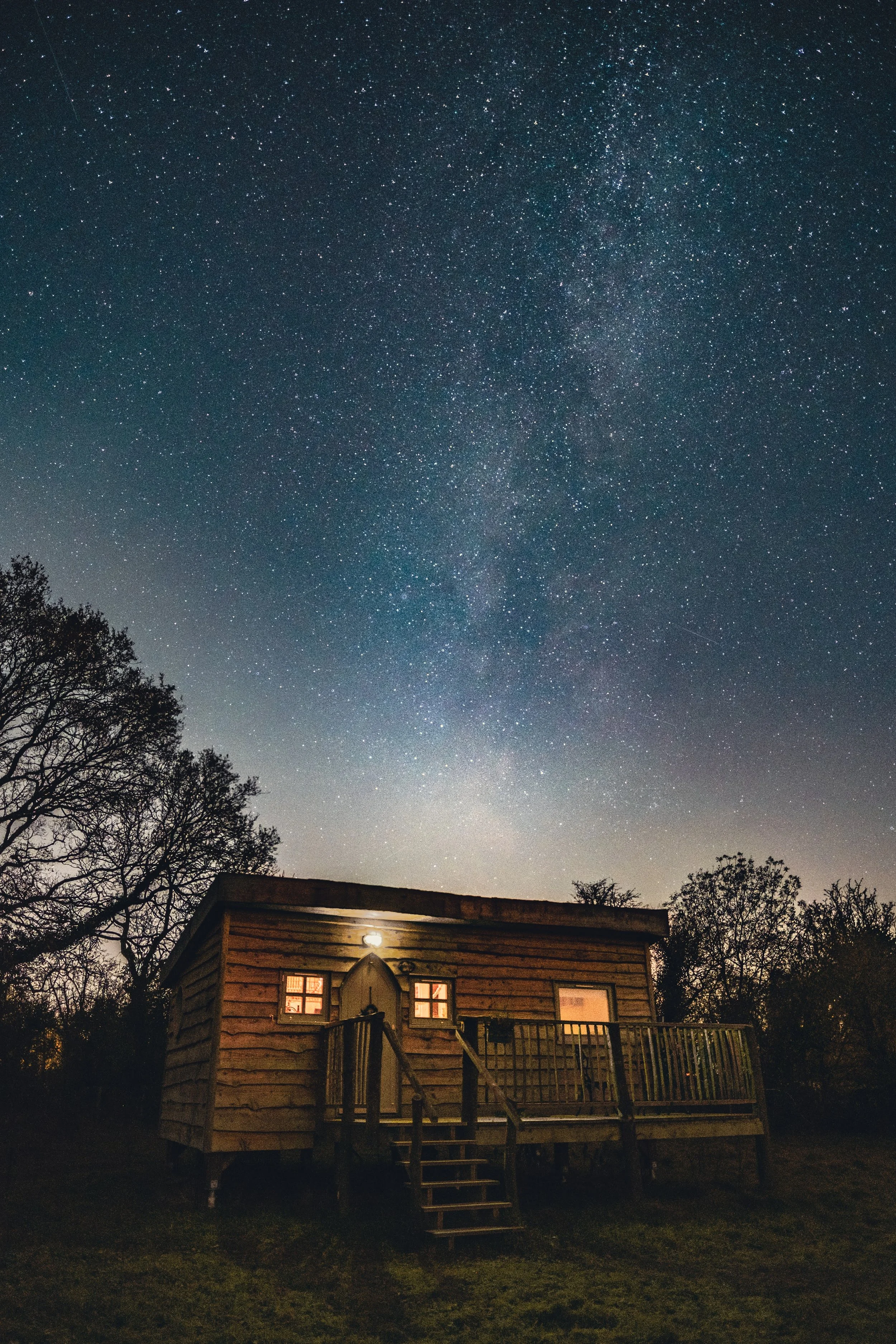 A small wooden cabin with lit windows and a porch, surrounded by trees, under a night sky filled with stars and the Milky Way galaxy.
