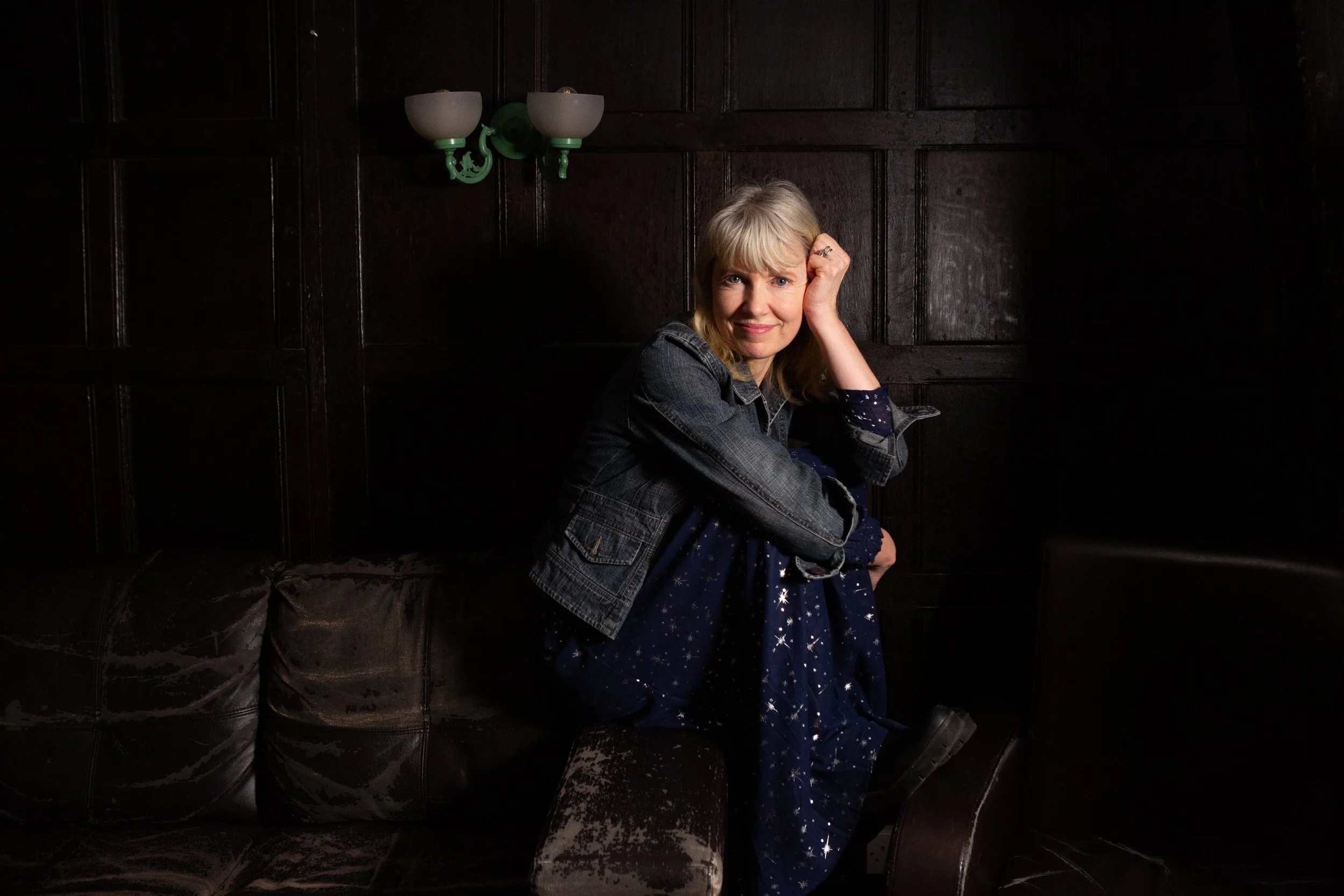 A woman with blonde hair sits on a dark brown leather couch, resting her head on her left hand, wearing a denim jacket and a navy dress with a star pattern, in a dimly lit room with dark wood paneling and a green wall sconce.