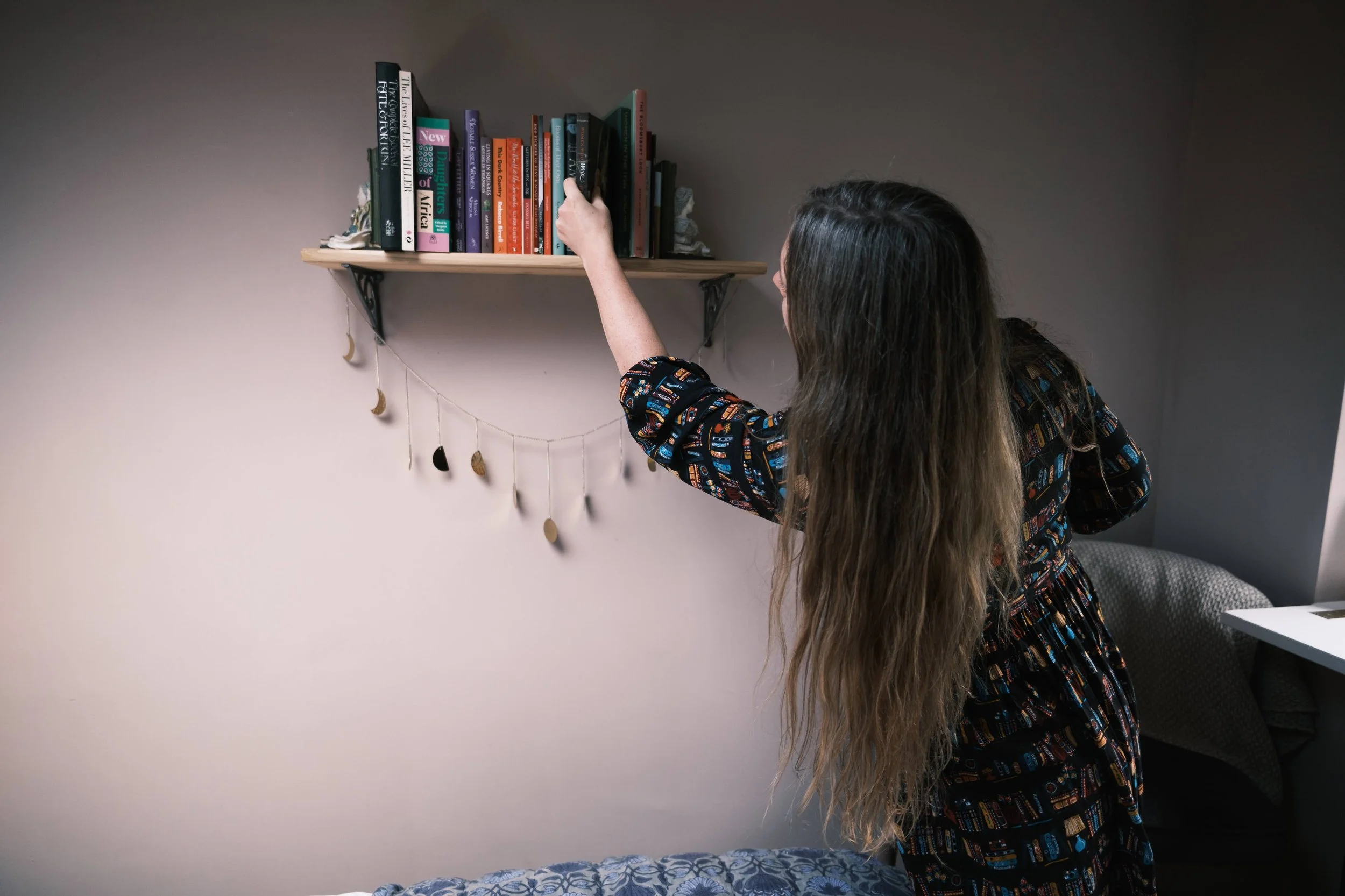 A woman with long brown hair reaching for a book on a bookshelf full of colorful books in a gray room.