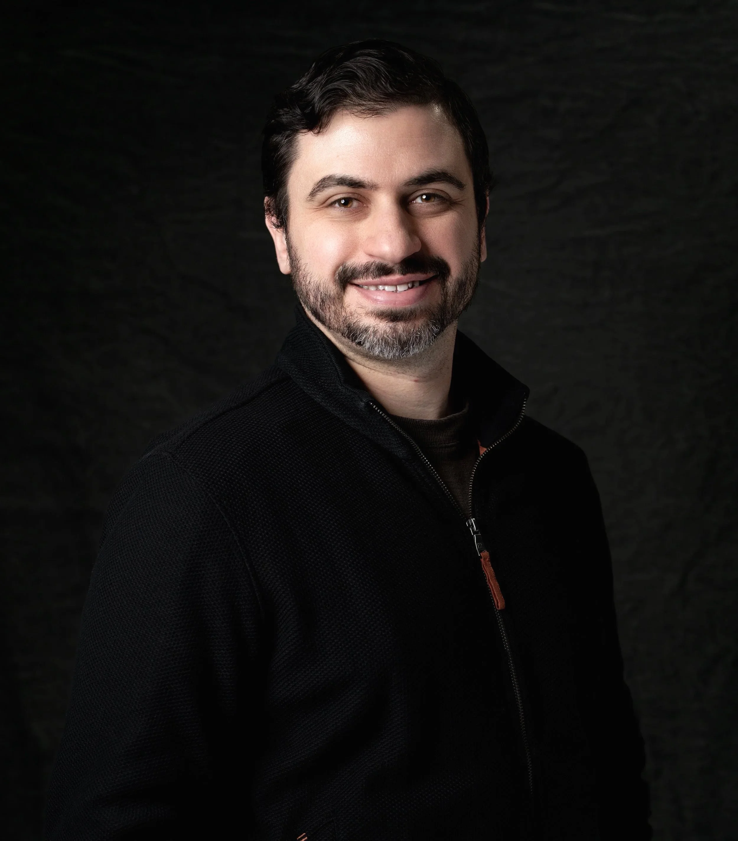 A man with dark hair, beard, and mustache smiling, wearing a black zip-up jacket, against a dark background.
