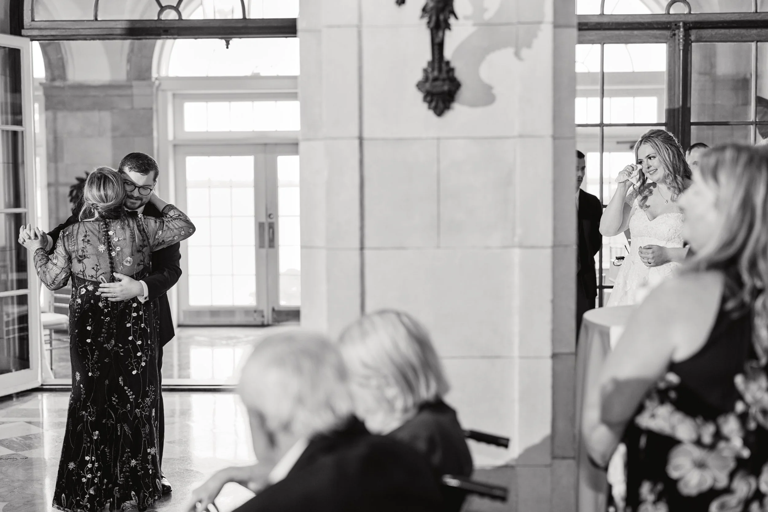 Black and white photo of a groom dancing with his mom. The bride is drying tears from her eyes. A pillar is in the center of the photo showing the groom and his mother on the left side and the bride on the right side of the photo.