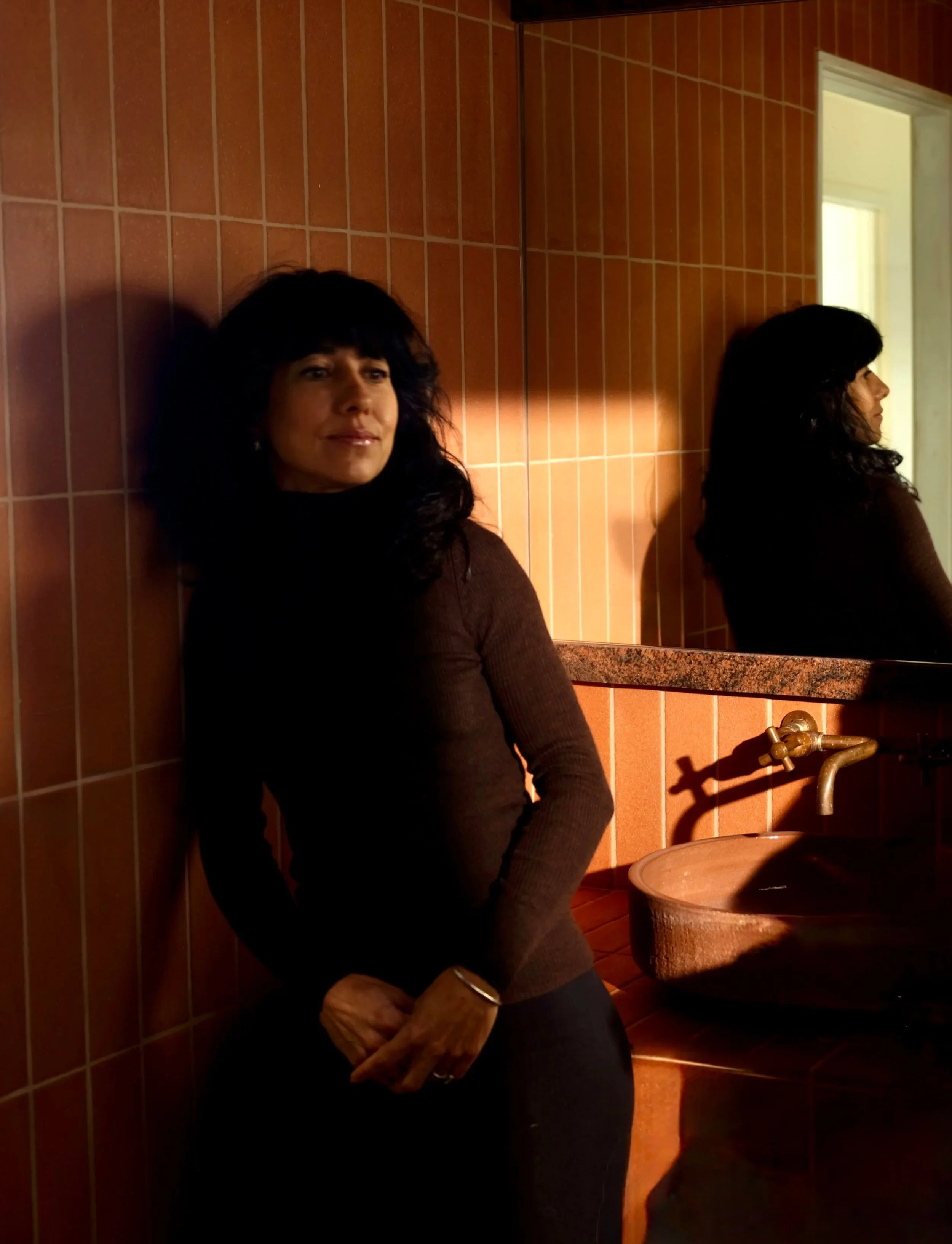 A woman with dark, curly hair wearing a long-sleeved, dark brown top, sitting against a tiled wall in front of a mirror, holding her hand in her lap, in a room with warm lighting and a rustic sink.