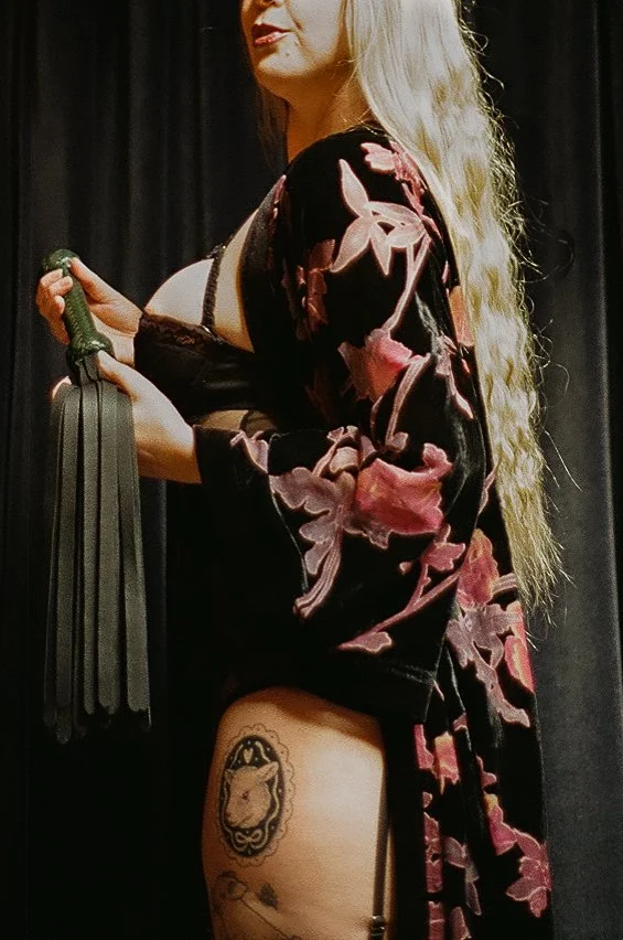 A woman with long, curly blonde hair wearing a kimono-style robe with floral patterns, holding a whip, with black curtains in the background.